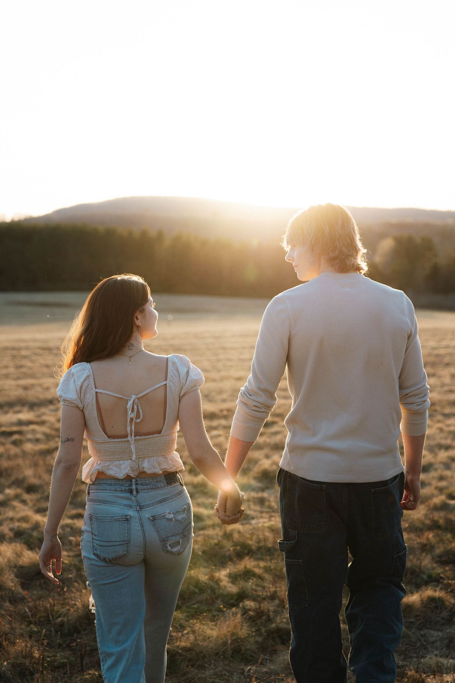 Couple walking hand in hand toward a radiant golden sunset in an open field.