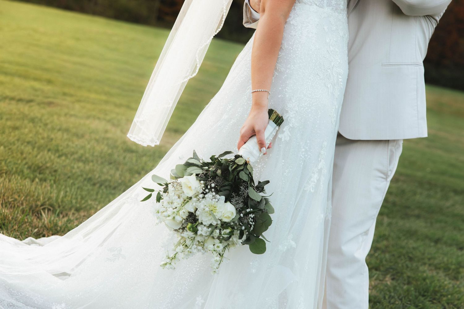 Wedding couple in white attire holding an elegant bouquet of white roses and greenery on grass.