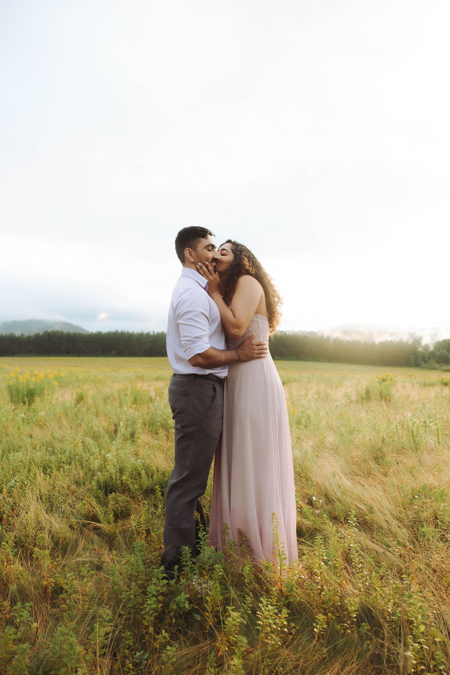 Couple embracing and kissing in a sunlit meadow, woman in pink flowing dress, mountains in background.