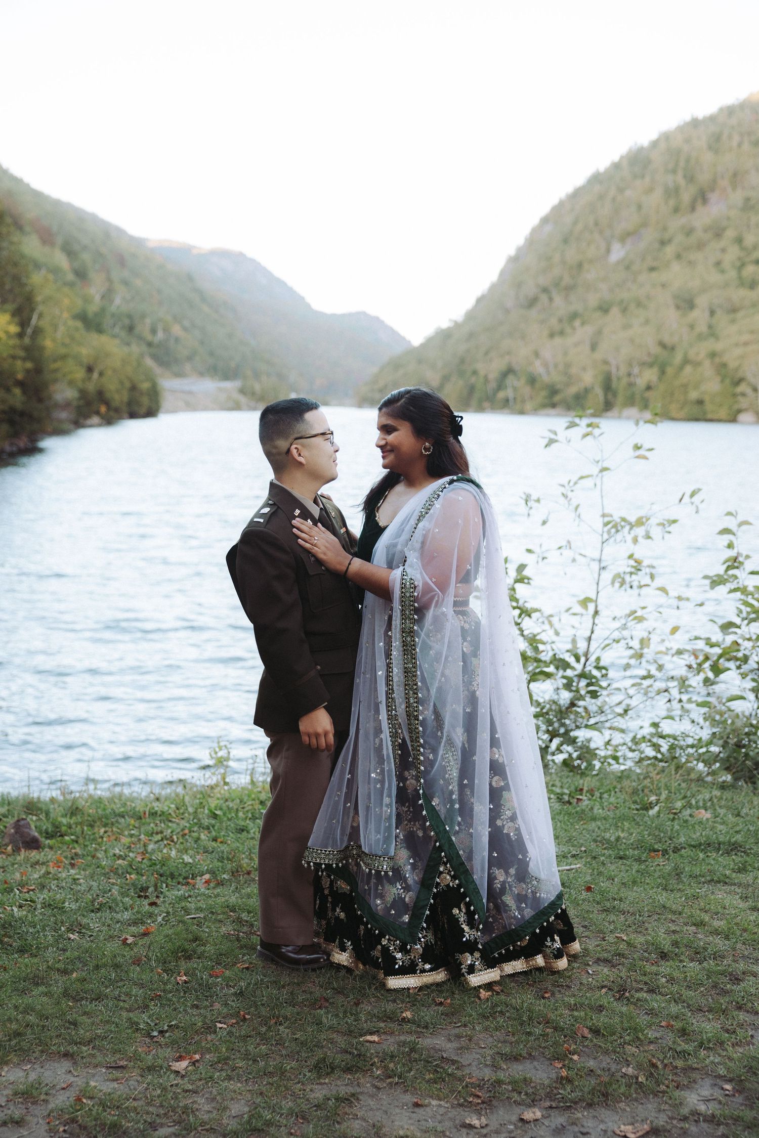 Couple in wedding attire smiling at each other by a mountain lake, wearing a suit and embroidered sari.