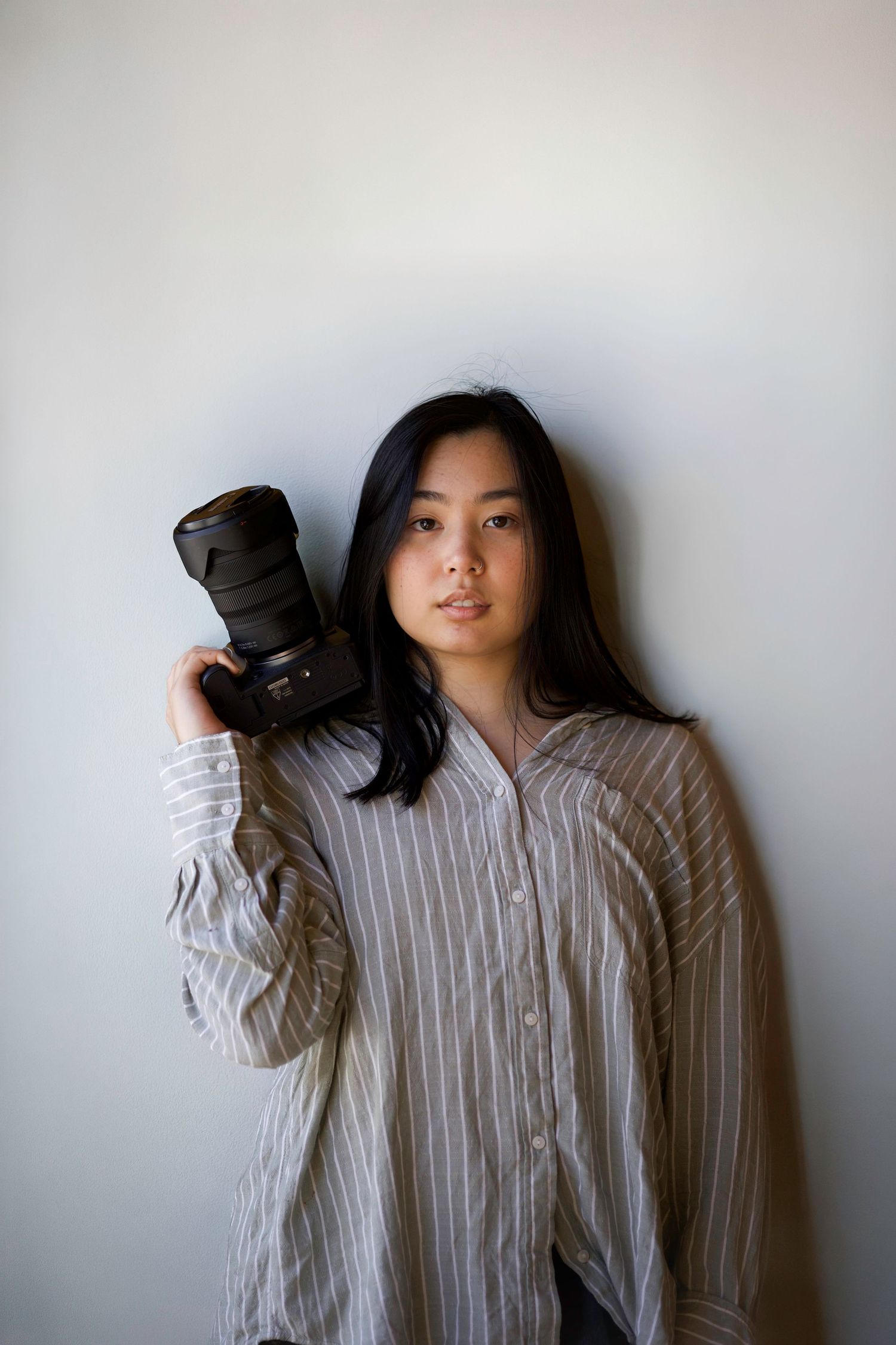 Young Asian woman photographer holding a professional DSLR camera on her shoulder against a white wall.