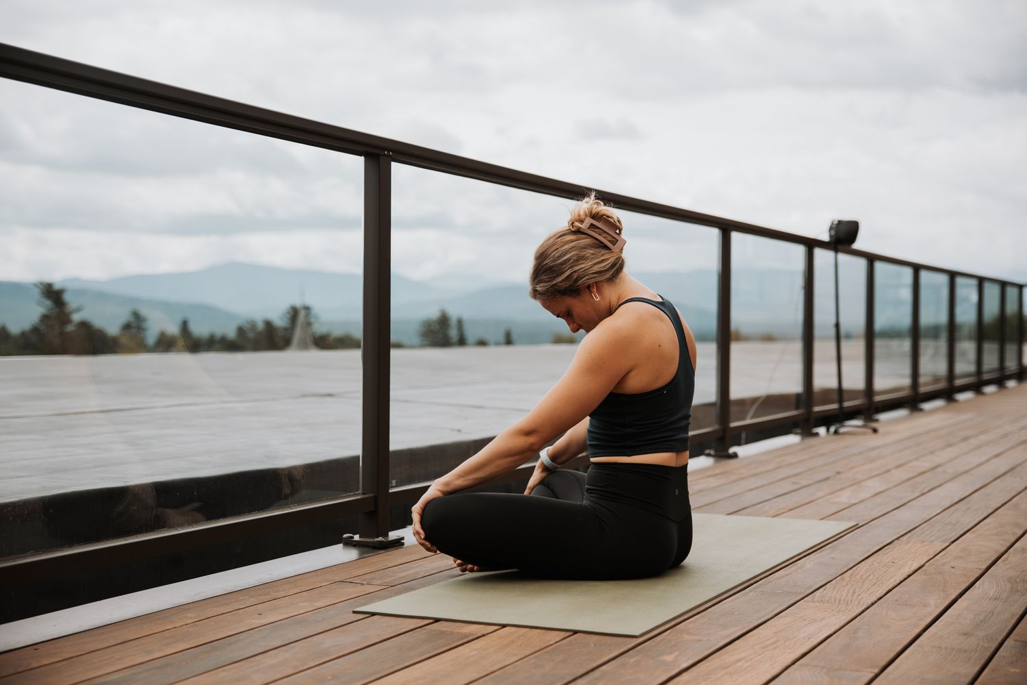 Woman in black activewear practicing yoga in seated pose on deck with mountain view.