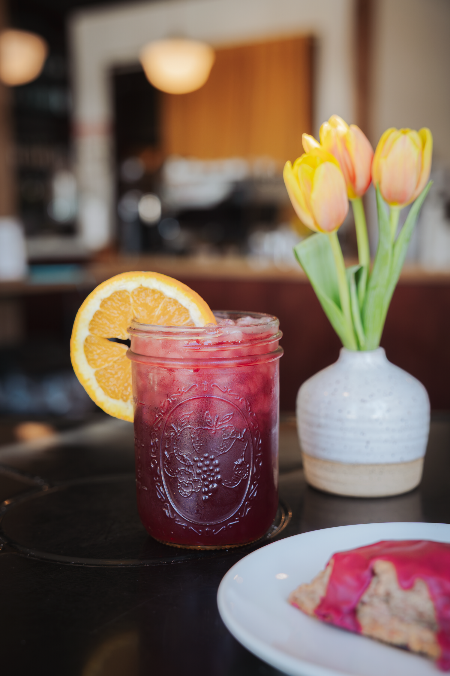Red berry iced drink in mason jar with orange slice beside tulips and dessert plate.