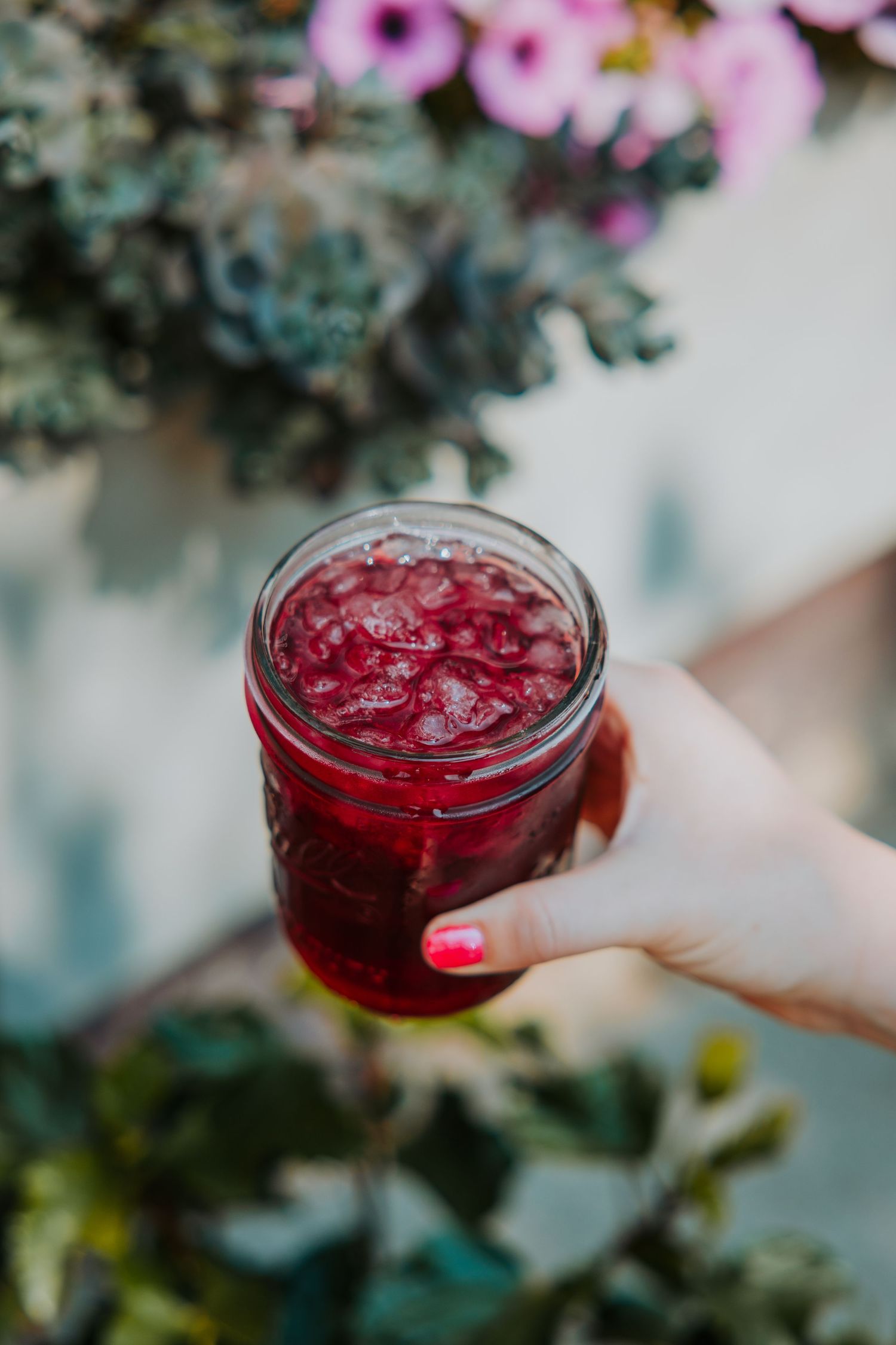 Hand holding a mason jar filled with a vibrant red berry drink outdoors near flowers.