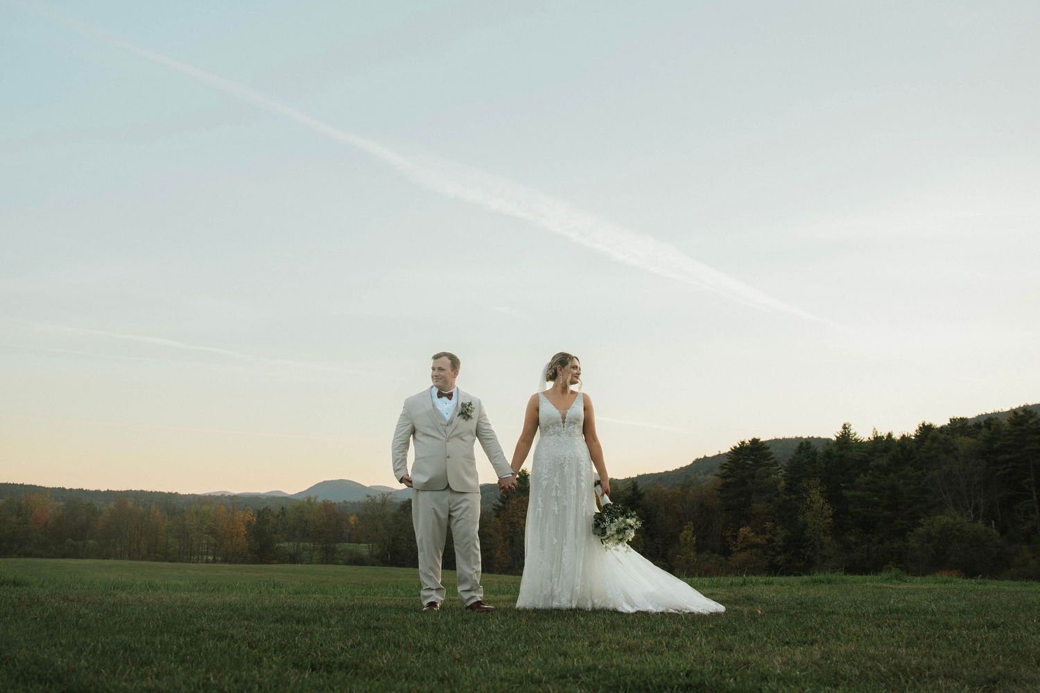 Bride and groom holding hands in open green field at sunset with mountain backdrop.