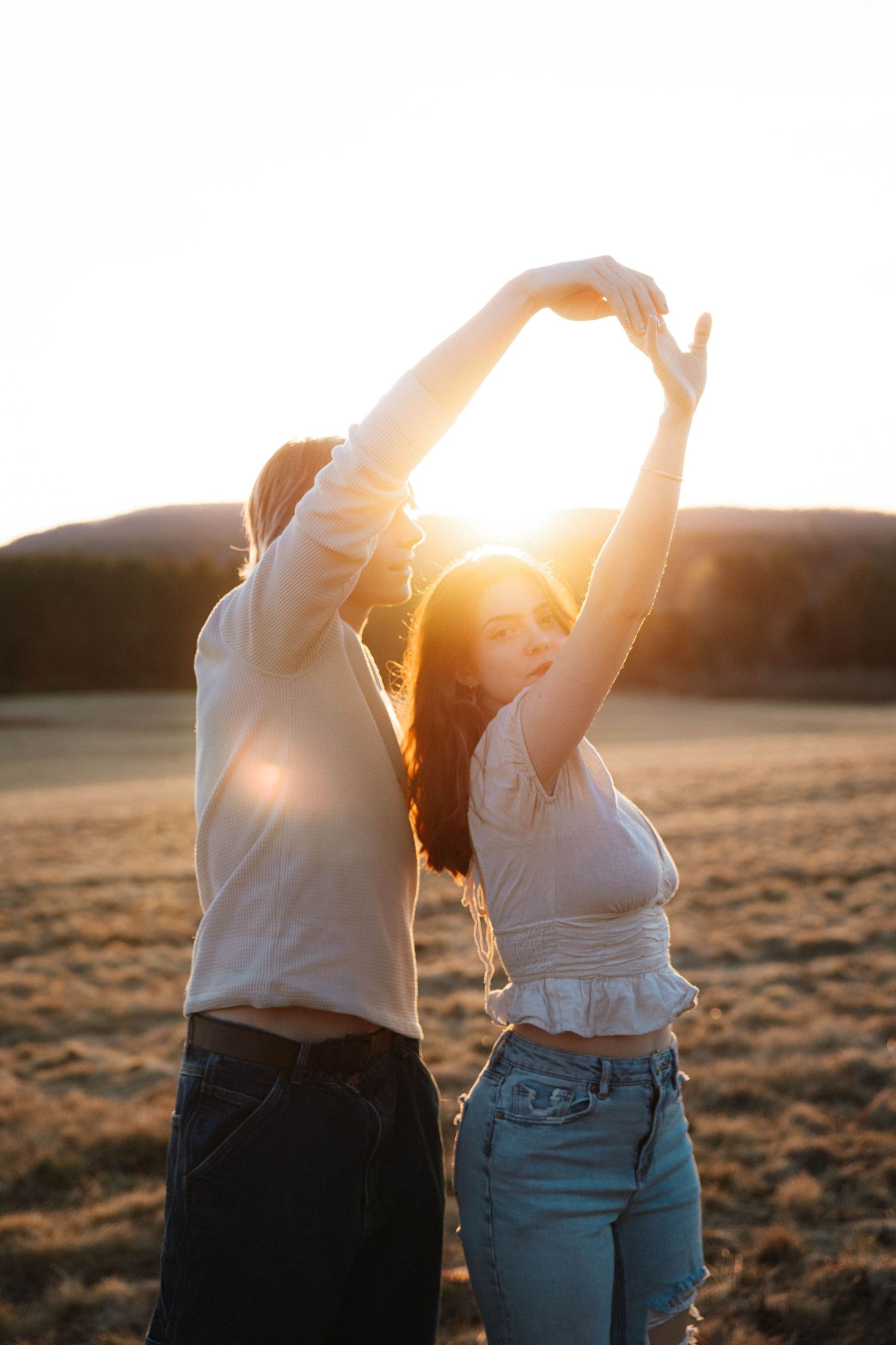 Two young women dancing together in a golden sunset field, raising their arms to form a heart shape against the glowing sky.