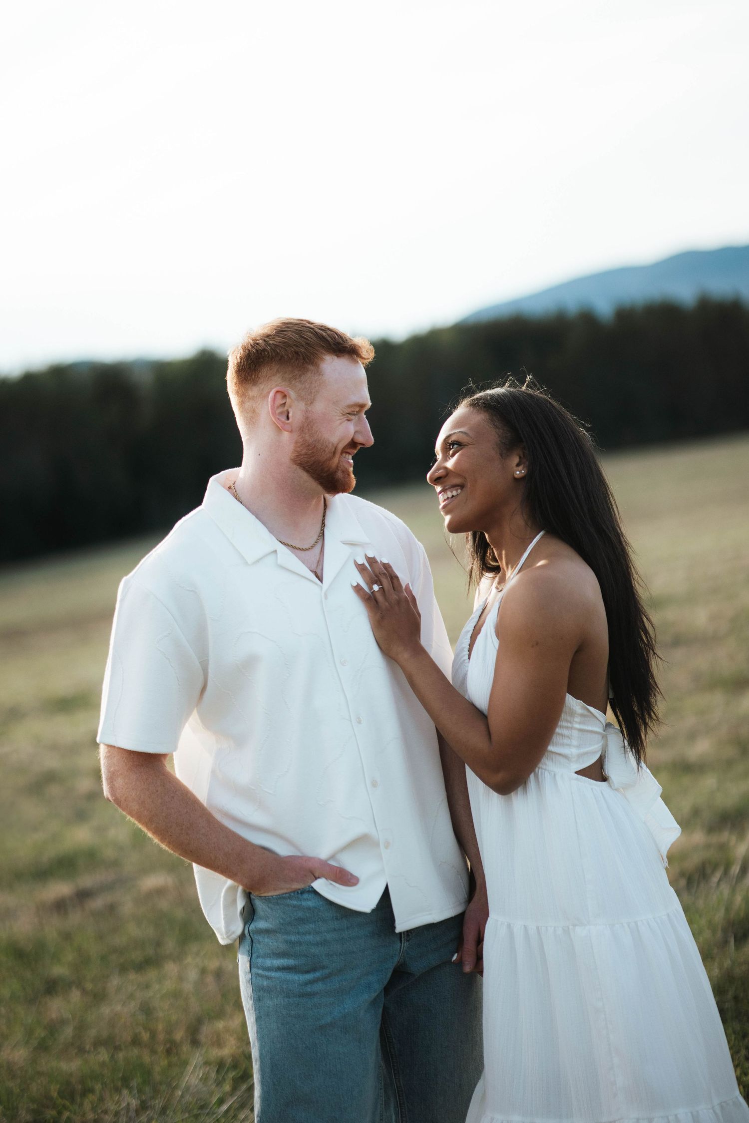 A smiling interracial couple in white outfits share a romantic moment in a scenic meadow with mountains in the background.