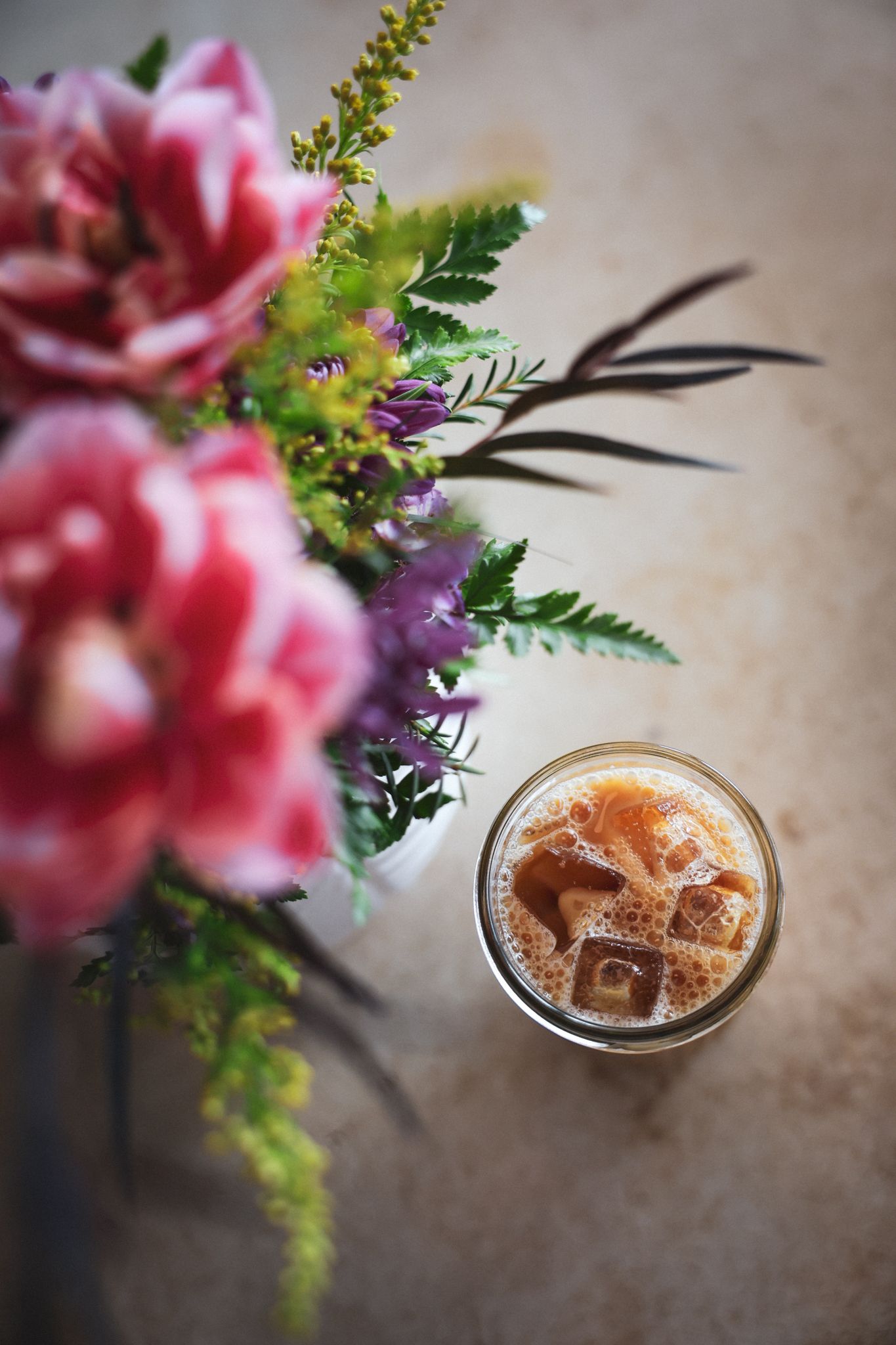 Overhead view of an iced coffee in a mason jar beside a pink floral bouquet on a table.