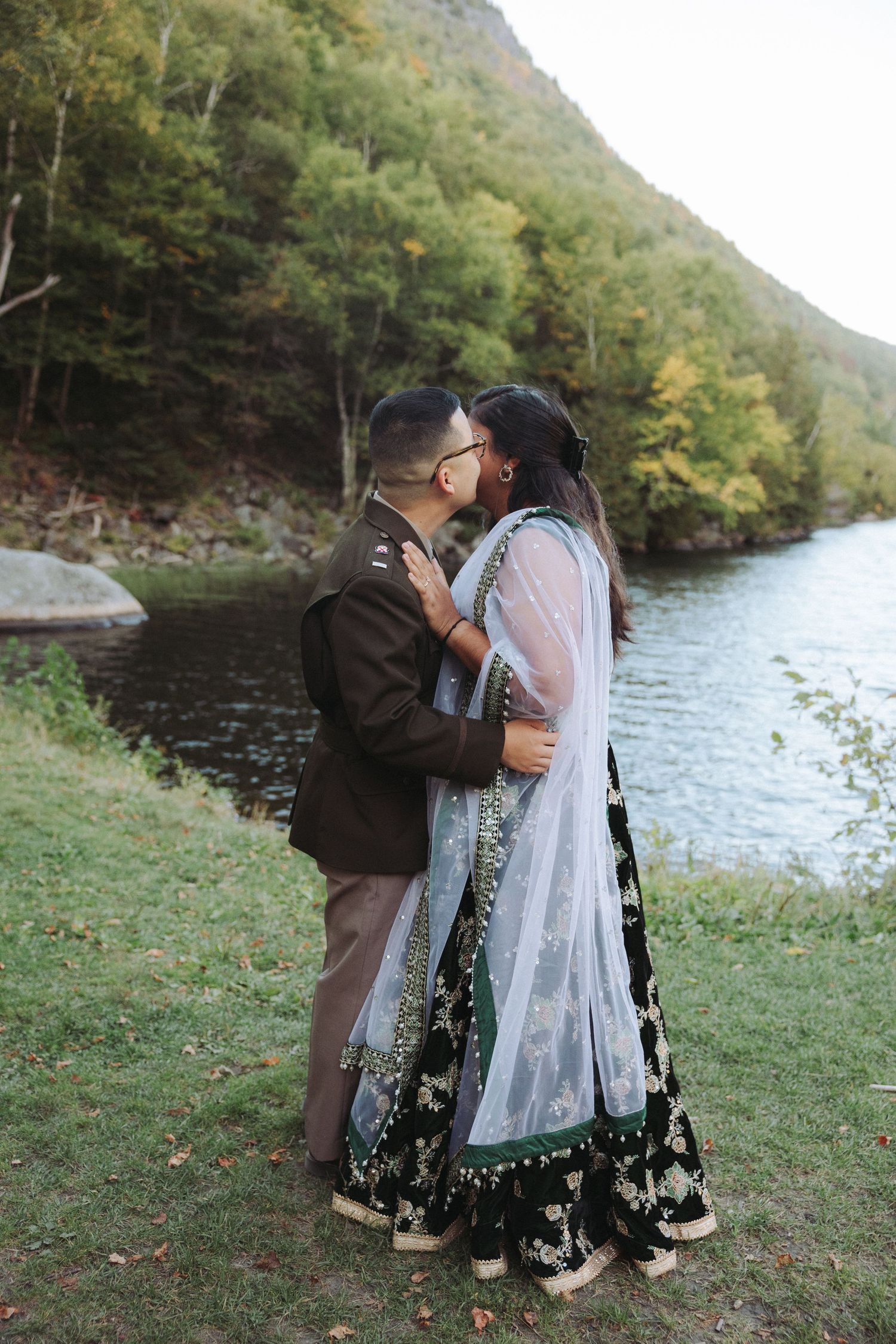 Couple sharing a kiss near a river, woman wearing traditional Indian lehenga with sheer dupatta.