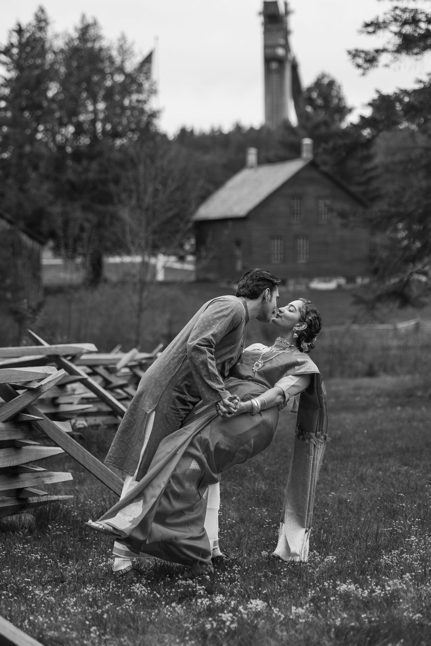Black and white photo of a couple sharing a romantic dip kiss outdoors near a rustic building and wooden fence.