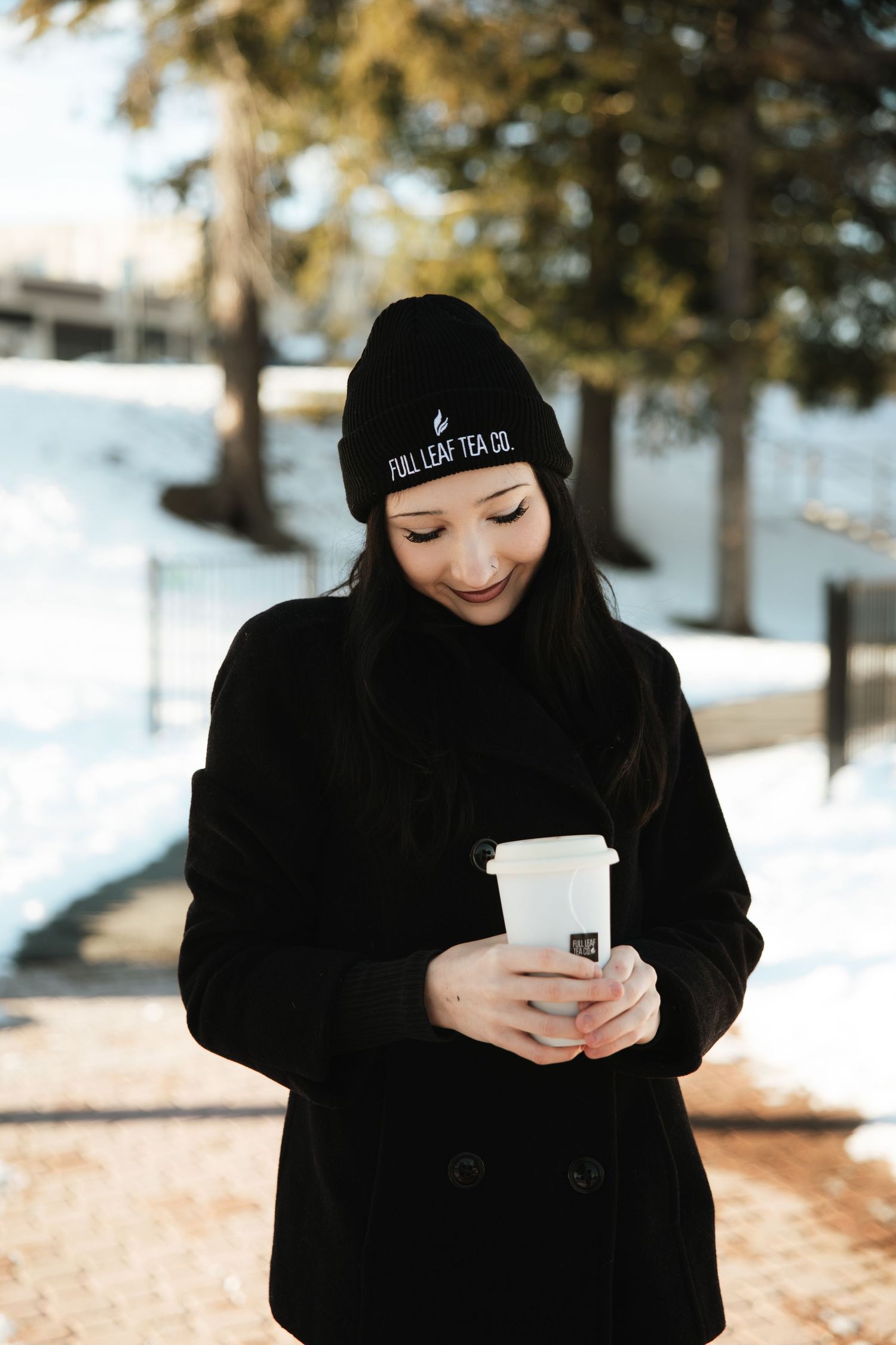 Smiling woman in black coat and Full Leaf Tea Co beanie holding a white cup in snow.