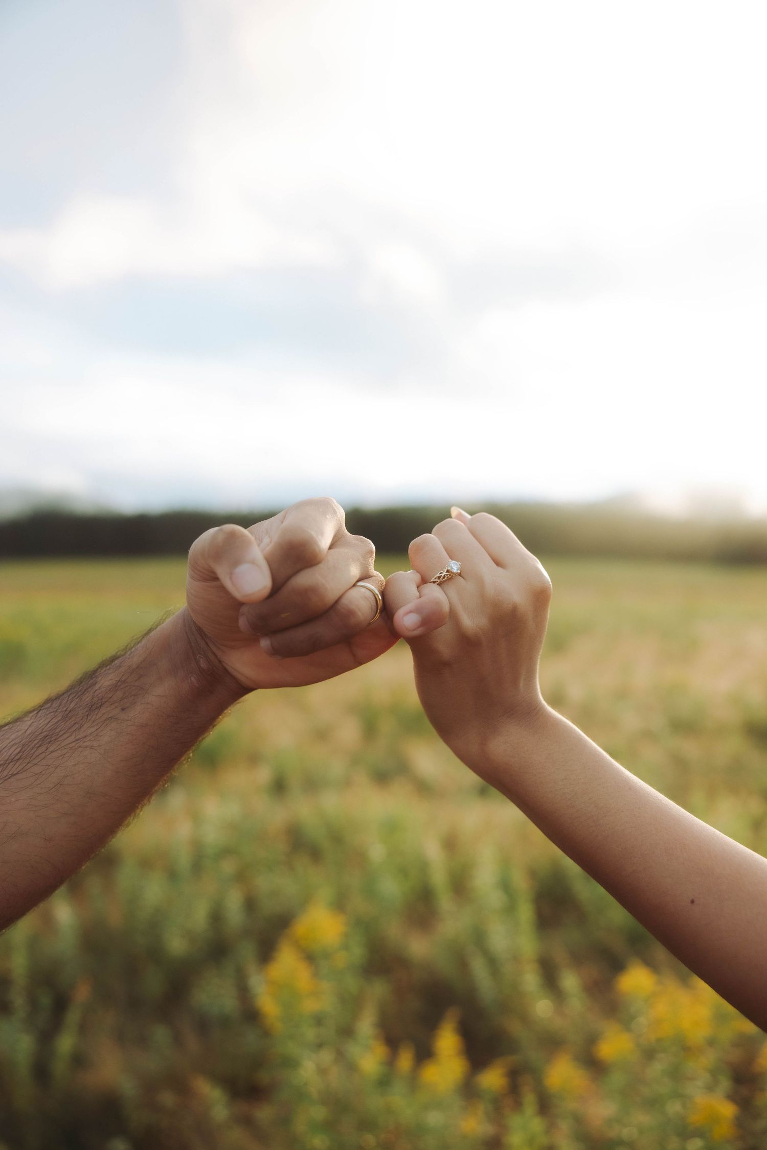 Close-up of couple doing a pinky promise with engagement ring visible in a field.