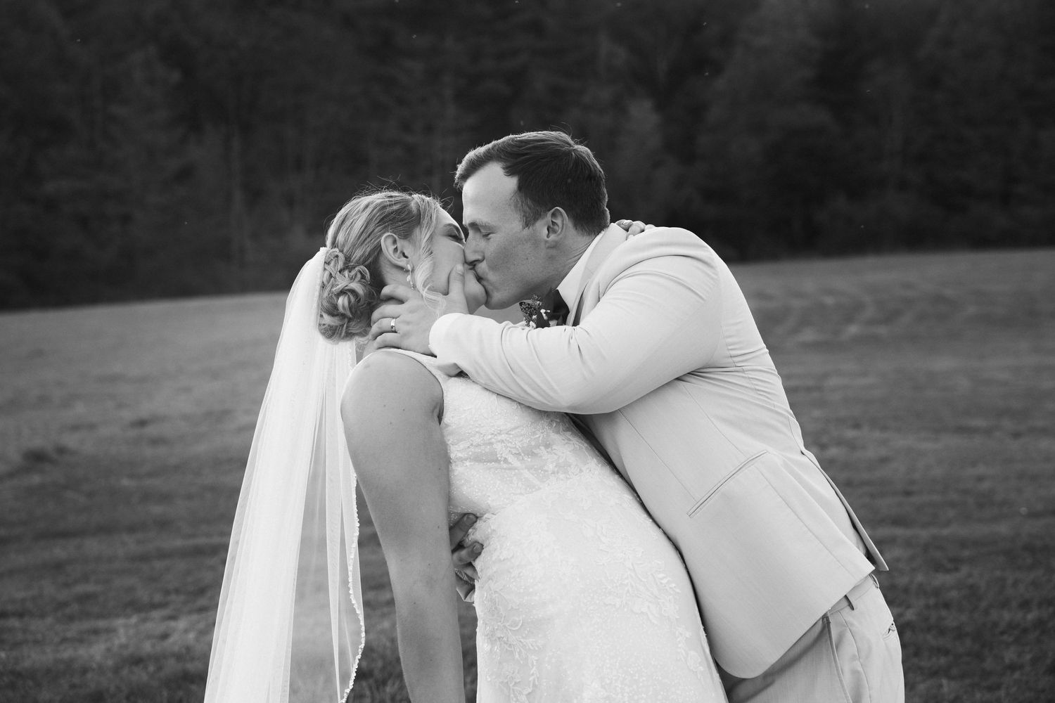 Bride and groom sharing romantic kiss in black and white wedding photo in scenic outdoor field.