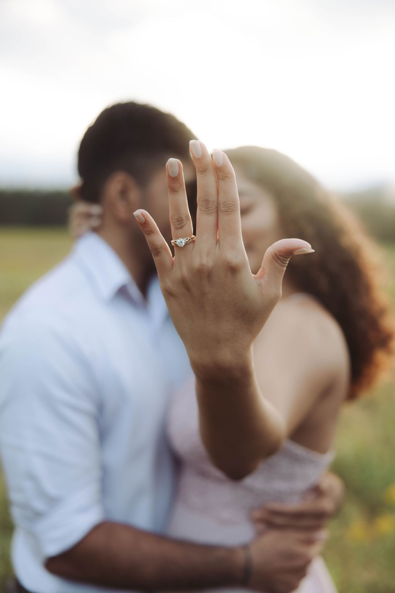 Engaged couple kissing outdoors while woman displays her diamond engagement ring to camera.