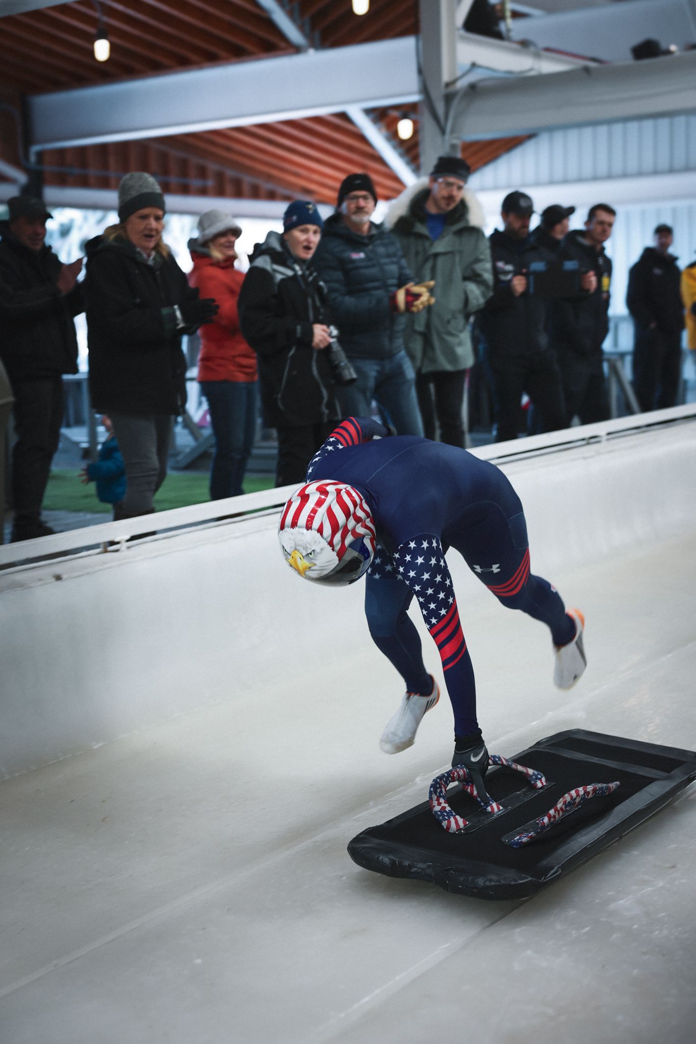 USA athlete in patriotic suit launches off skeleton sled at indoor ice track while spectators watch from sidelines.