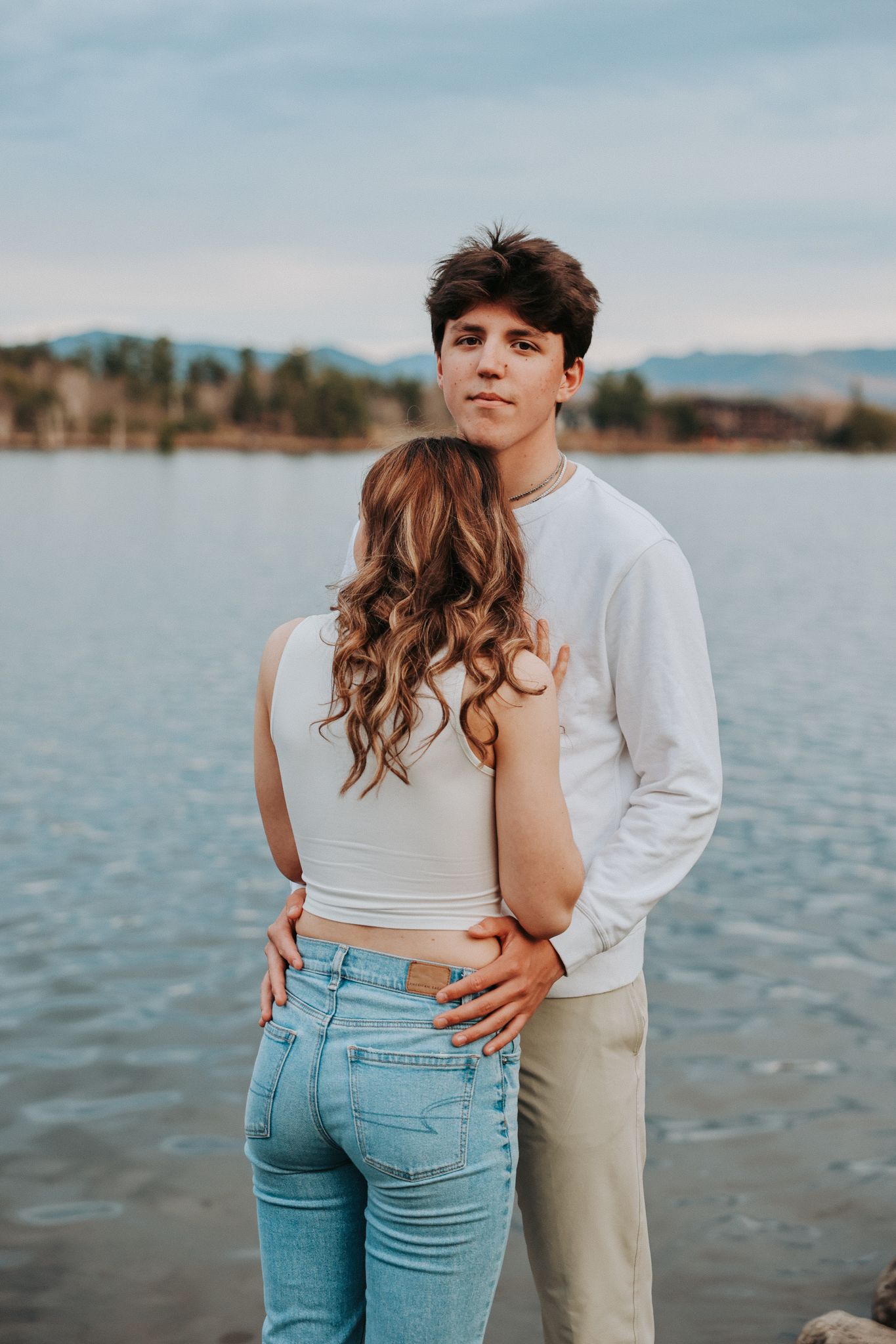 Young couple embracing by a lake, man in white sweatshirt holding woman in jeans from behind.