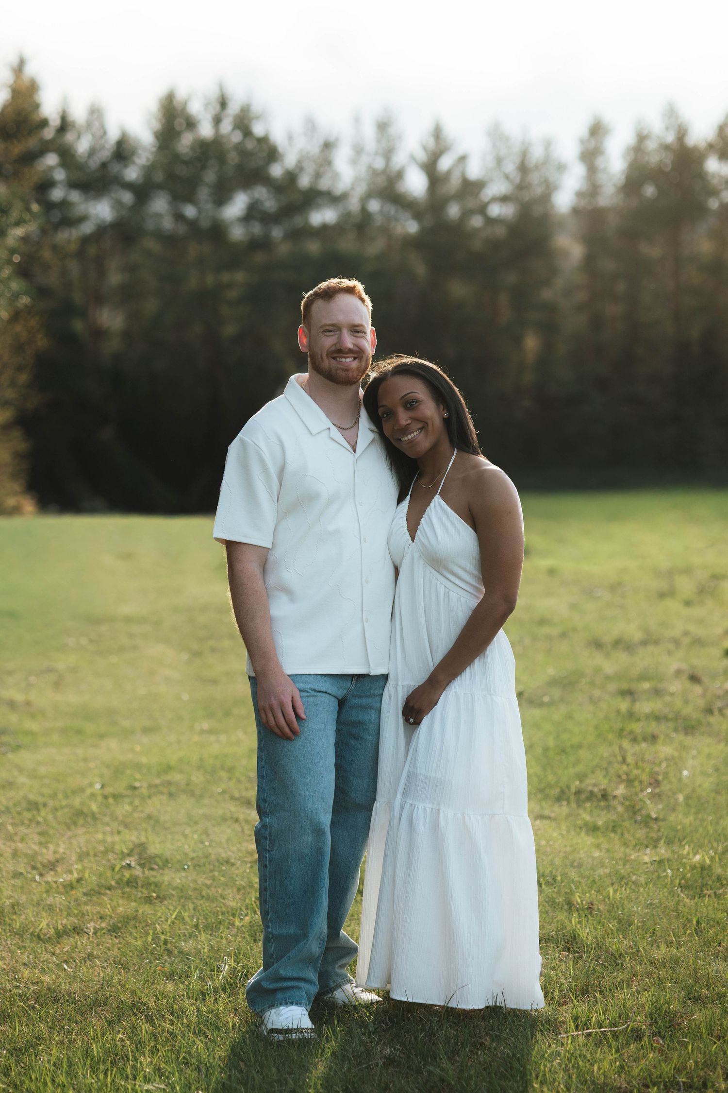 A smiling interracial couple in matching white outfits stands together on a green lawn with tall trees in the background.
