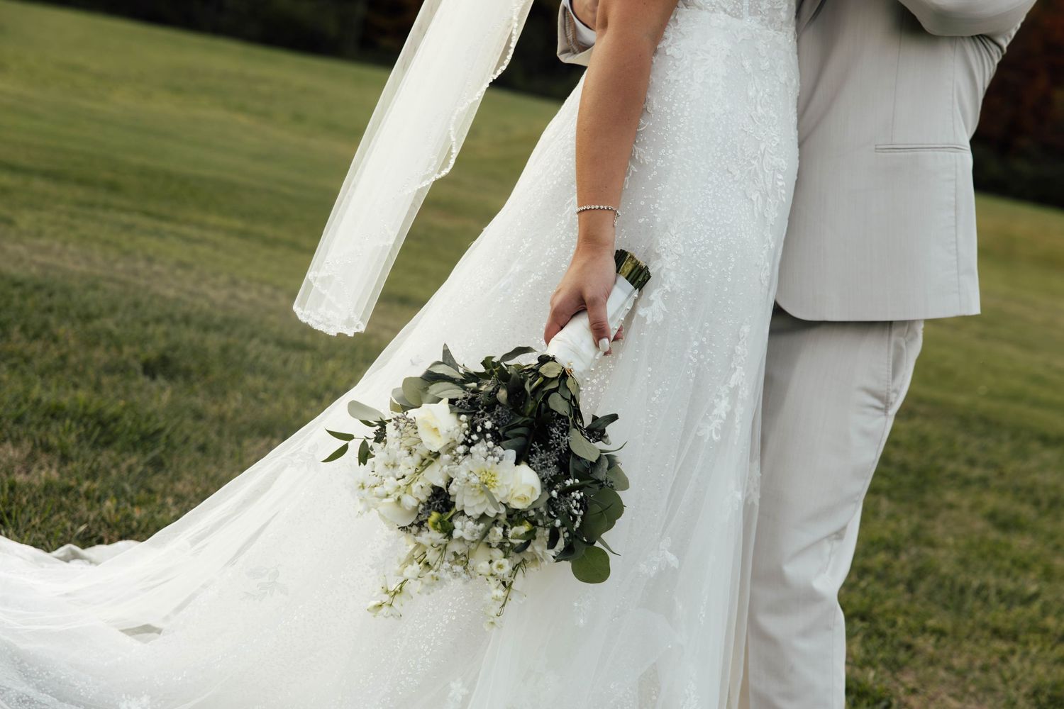 Bride in white lace gown holding elegant bouquet of white flowers and greenery stands with groom in light gray suit.