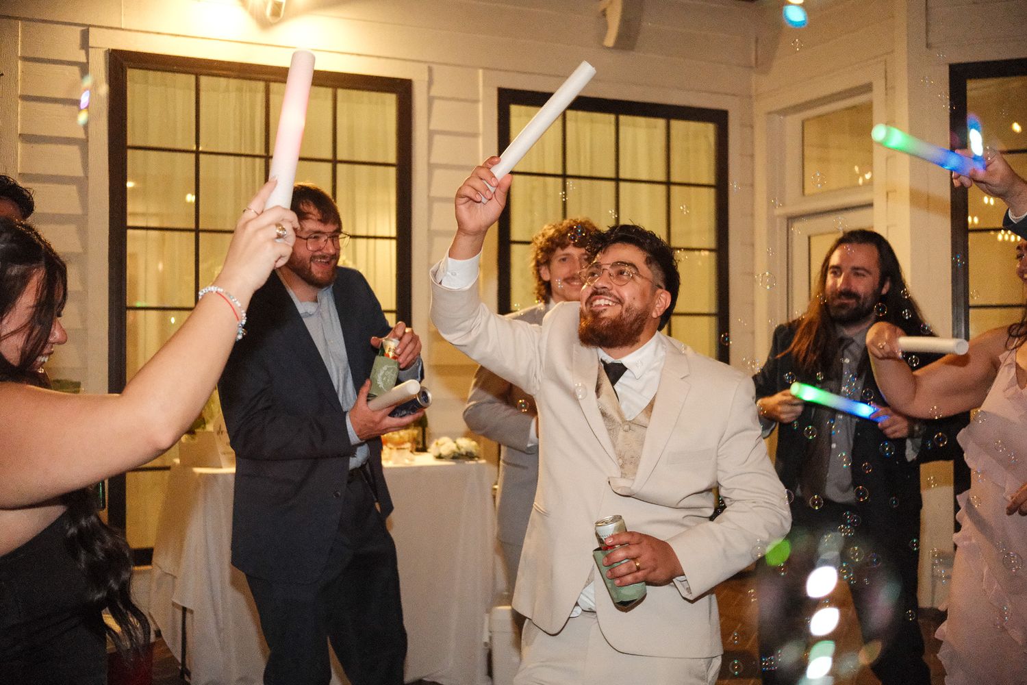 Groom in white suit celebrates at wedding reception holding glow stick among guests.