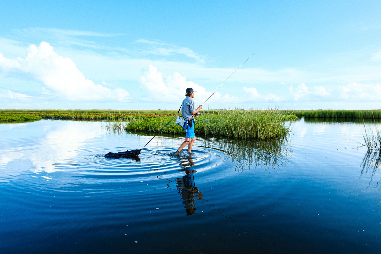 Man fishing on a paddleboard on calm blue water surrounded by marsh grass under blue sky.