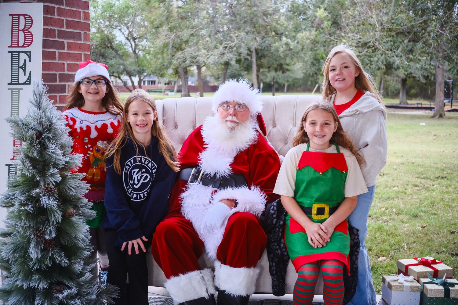 Four girls posing with Santa Claus outdoors near a Christmas tree and holiday decorations.