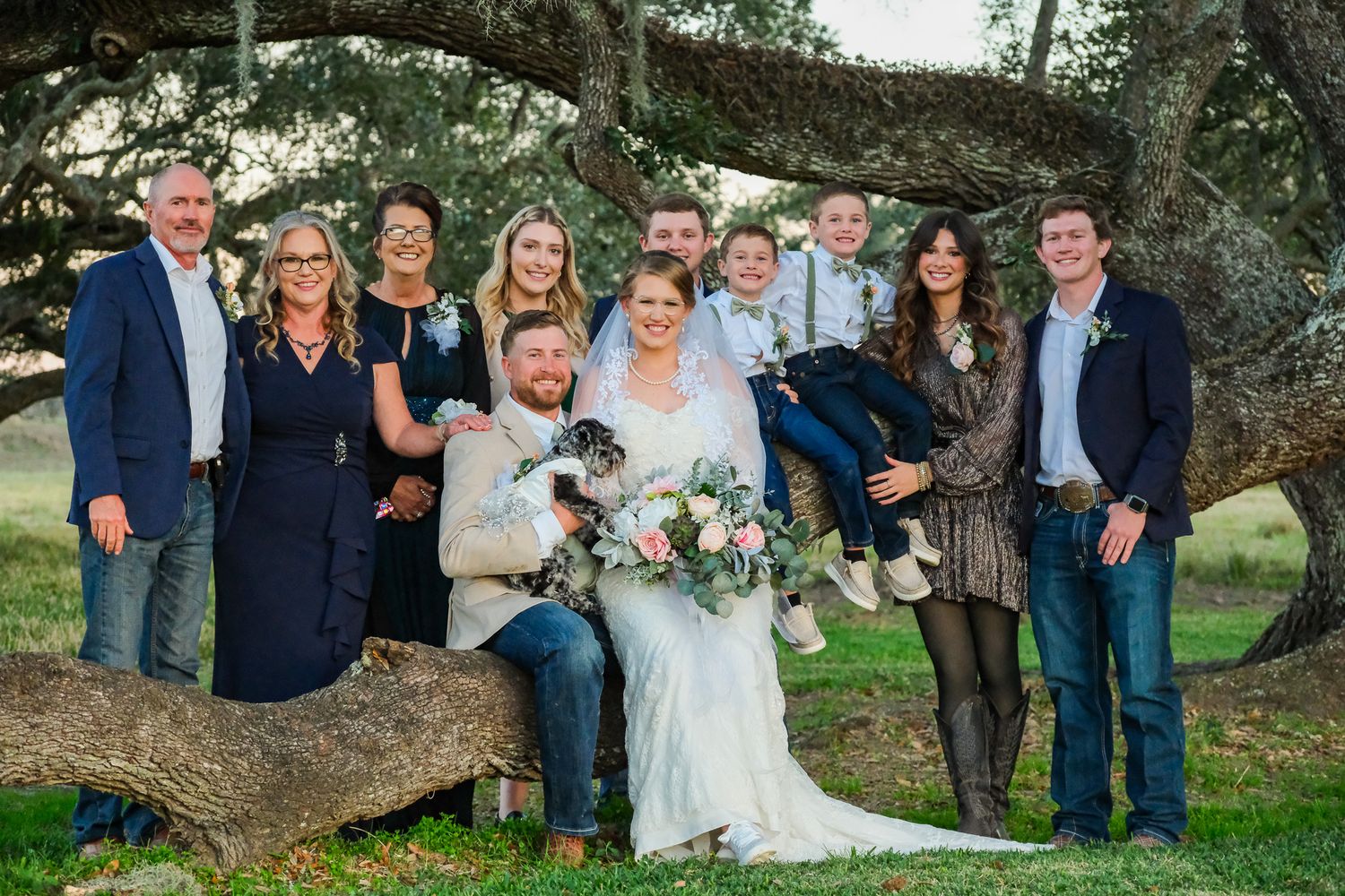 Wedding party poses outdoors under large oak tree, bride holding bouquet, groom seated, family gathered in formal attire.