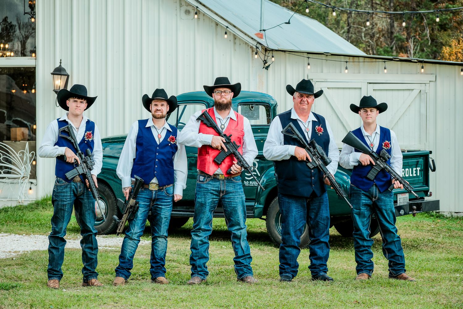 Five groomsmen in cowboy hats and vests pose with rifles in front of a vintage truck at a barn wedding.