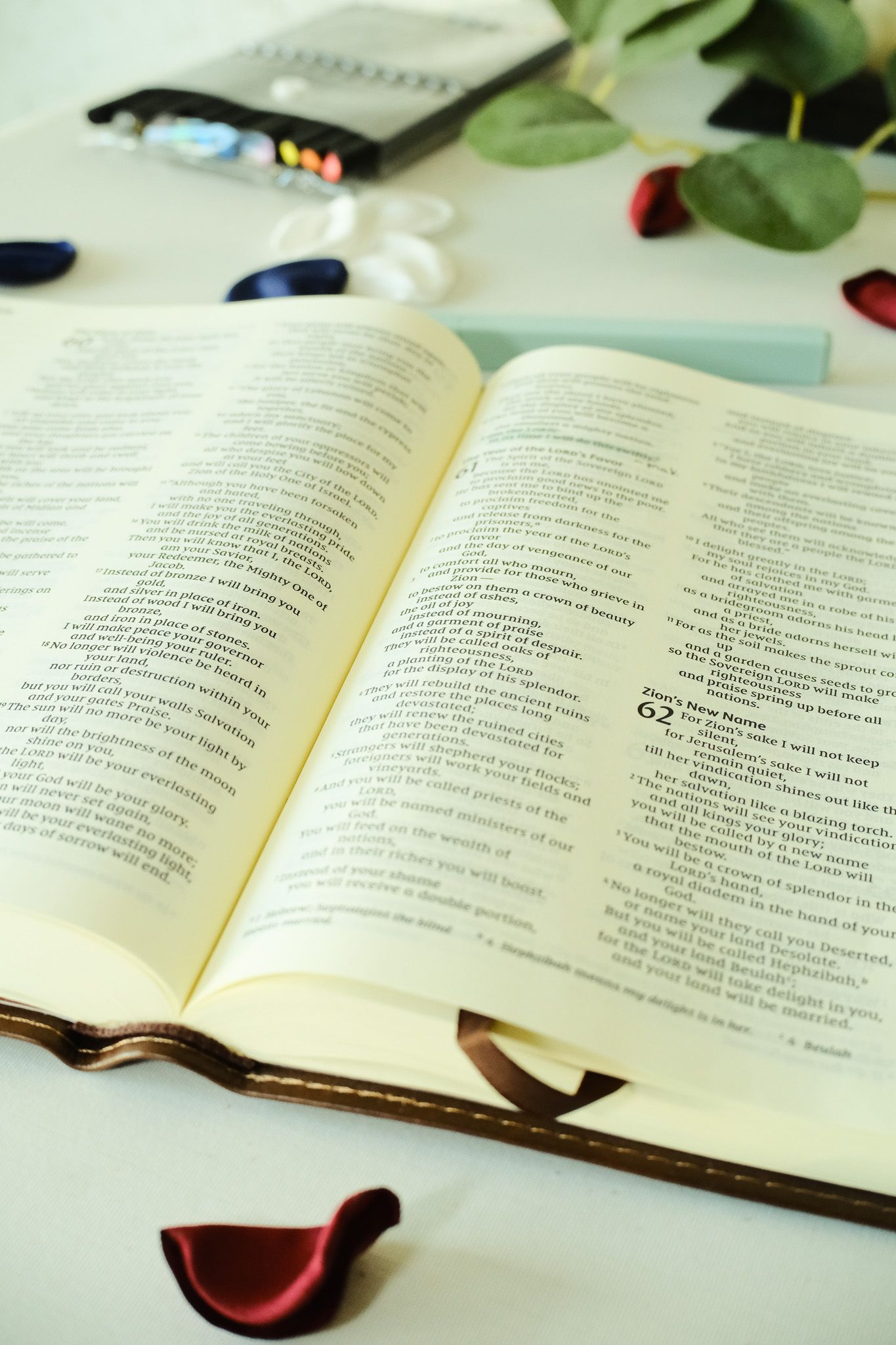 An open Bible with rose petals, ribbon bookmarks, and glasses resting on a white surface for a wedding ceremony.