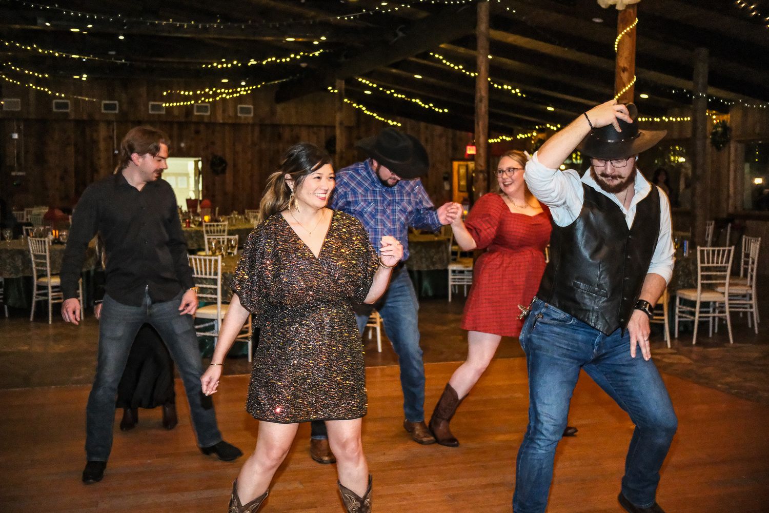 Two couples line dancing together in a rustic barn wedding reception venue with string lights.