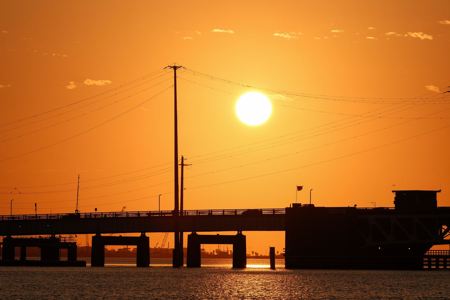 Dramatic orange sunset silhouetting a bridge and utility poles over calm water.
