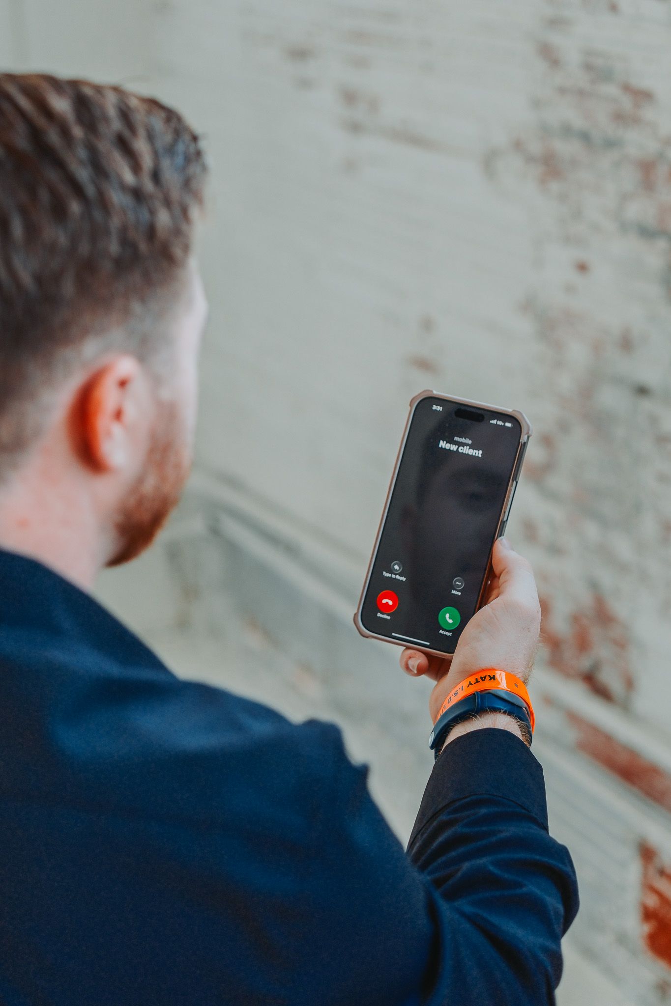 Man in navy jacket views incoming call on iPhone with orange wristband visible.
