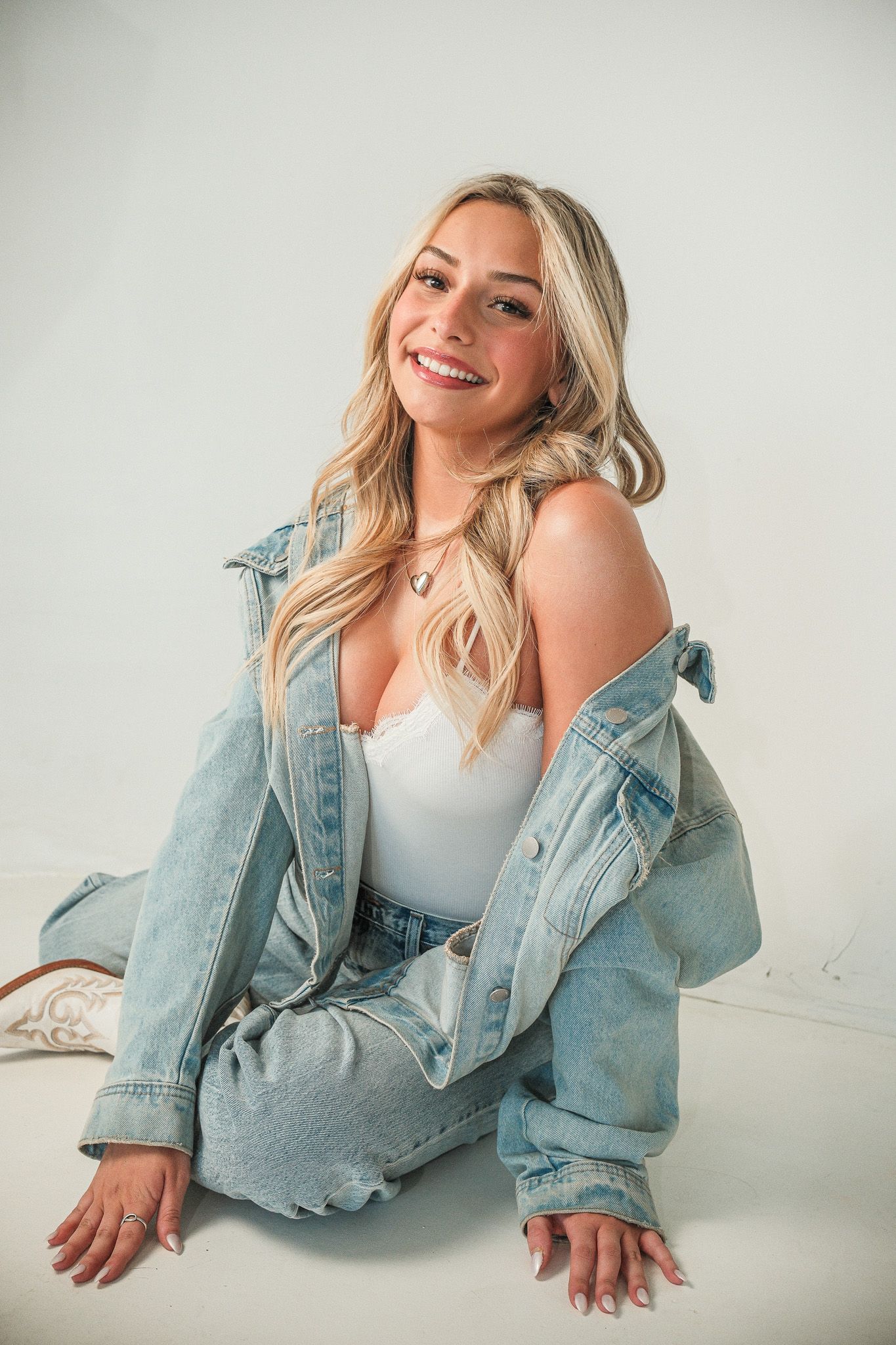 Smiling blonde woman in a white top and denim jacket sitting on a white studio floor.