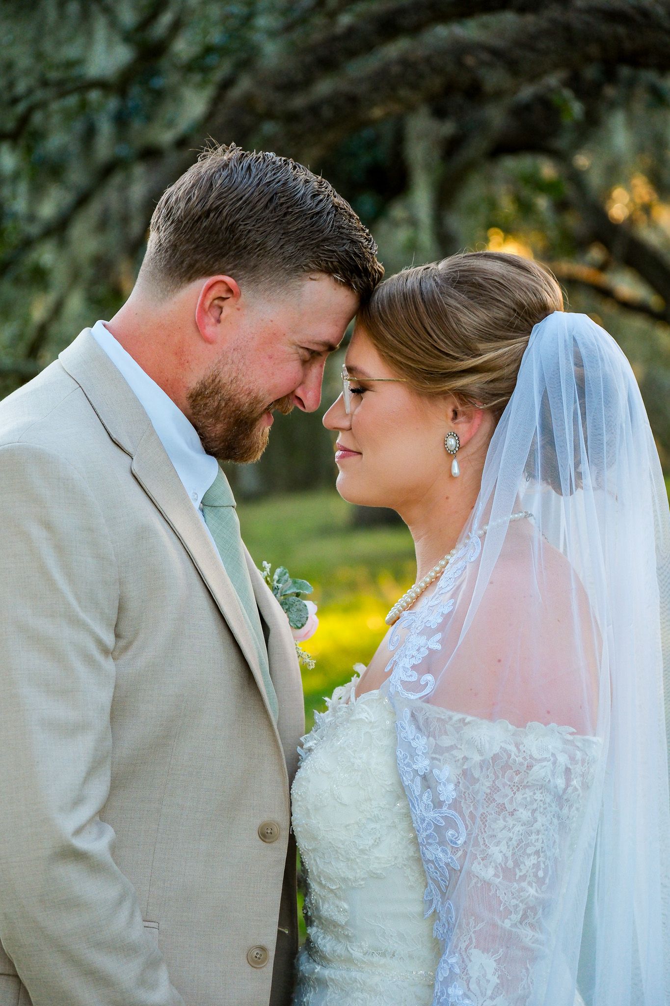 Romantic close-up of bride and groom touching foreheads tenderly in golden hour light beneath oak trees.
