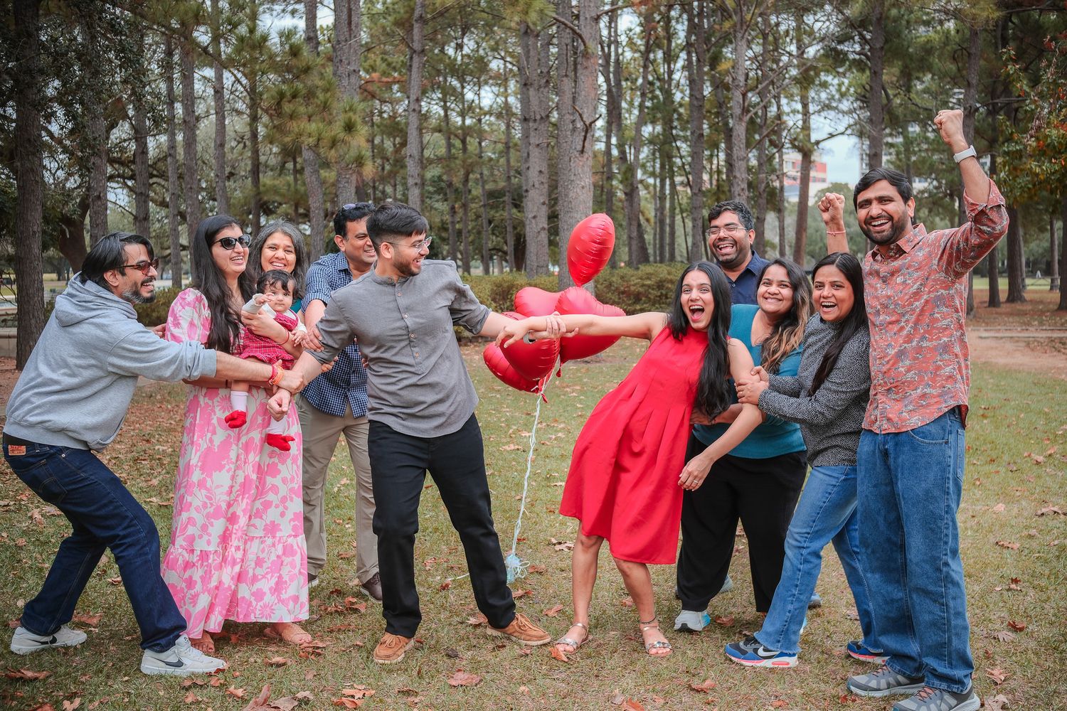 Group of friends joyfully posing outdoors in a park with pine trees, surrounding a woman in a red dress holding balloons.