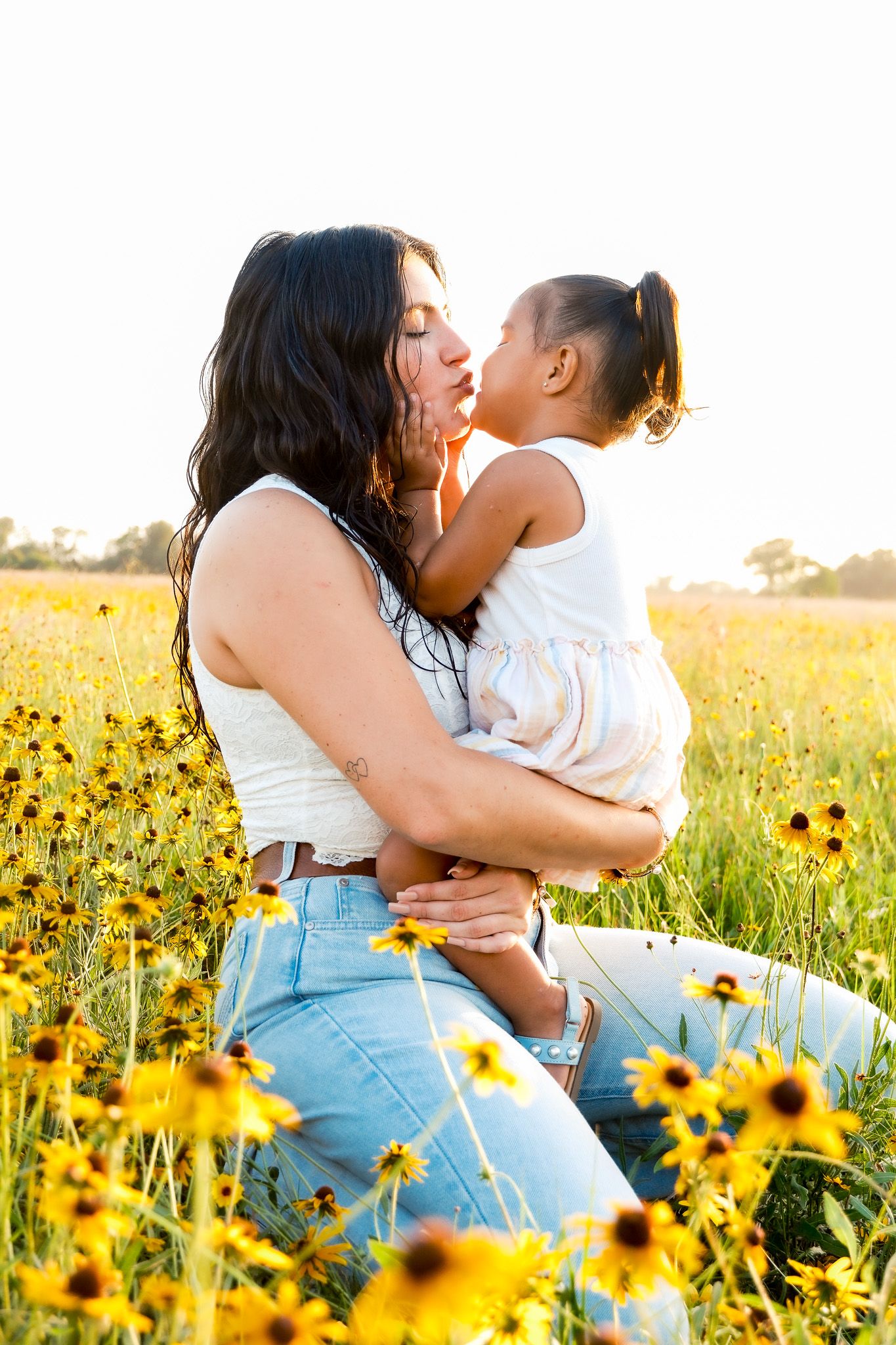 Mother and young daughter sharing a kiss in a sunny yellow wildflower field, wearing white tops and jeans.