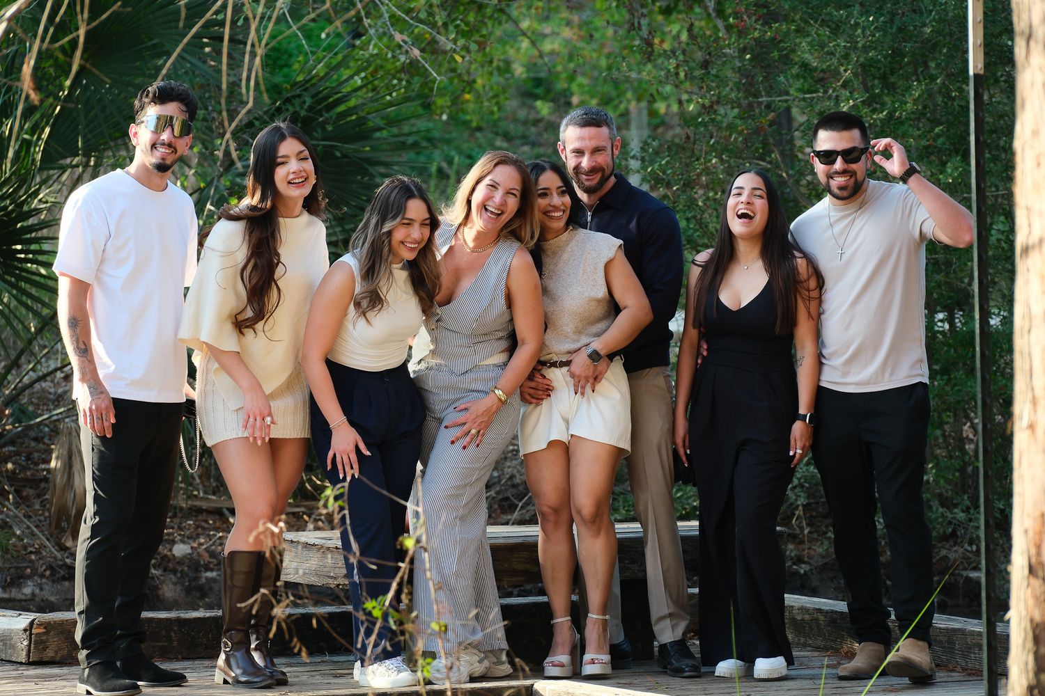 Group of seven friends smiling and laughing together outdoors in a lush green garden setting.