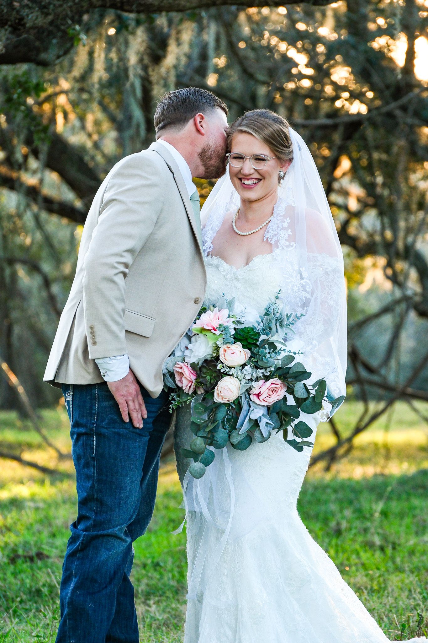 Groom in tan suit whispering to smiling bride holding pink and white floral bouquet in outdoor setting.