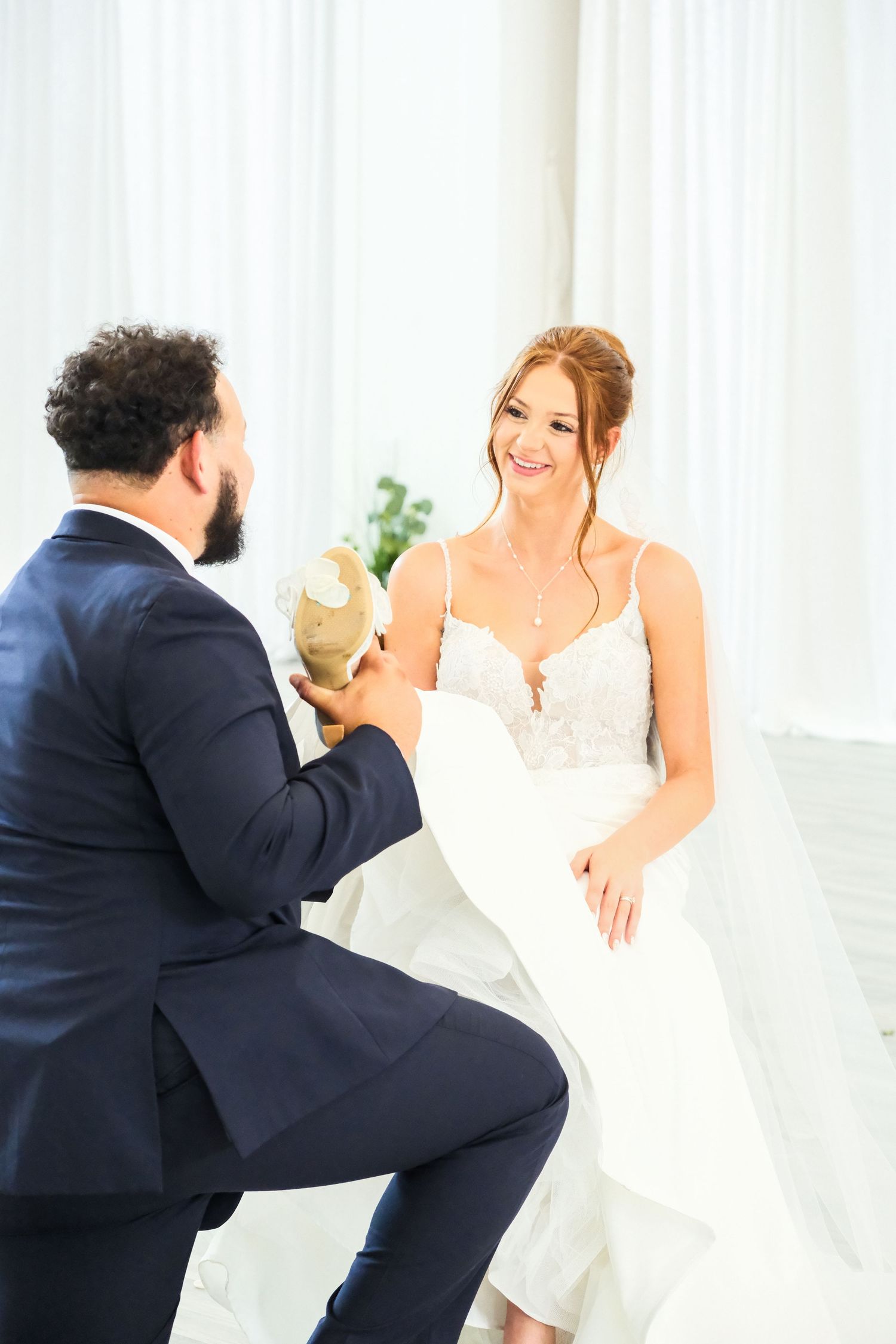 Groom kneeling to put shoes on smiling bride in white lace wedding dress in bright white room.