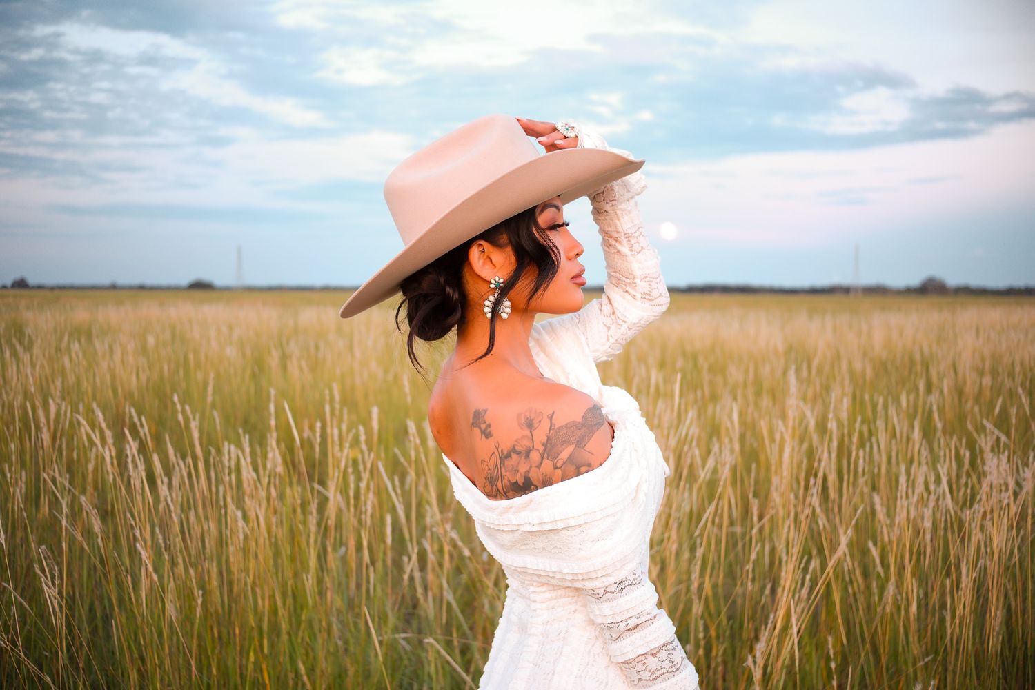 Woman in white lace dress and tan hat standing in tall golden grass field at dusk.