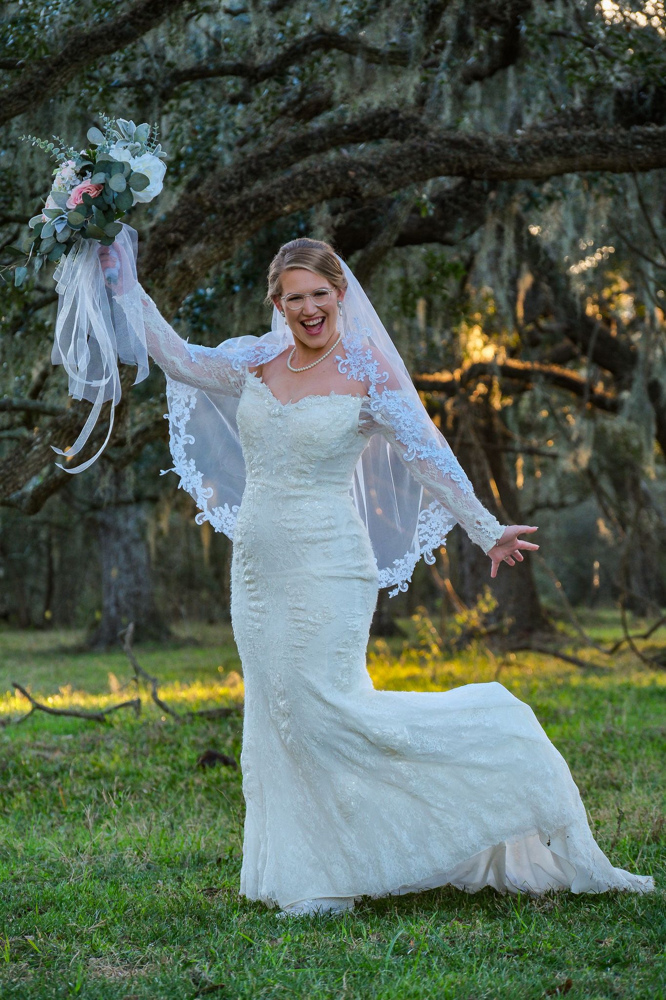 Joyful bride in lace mermaid gown and veil celebrating outdoors under moss-covered oak trees with bouquet raised.