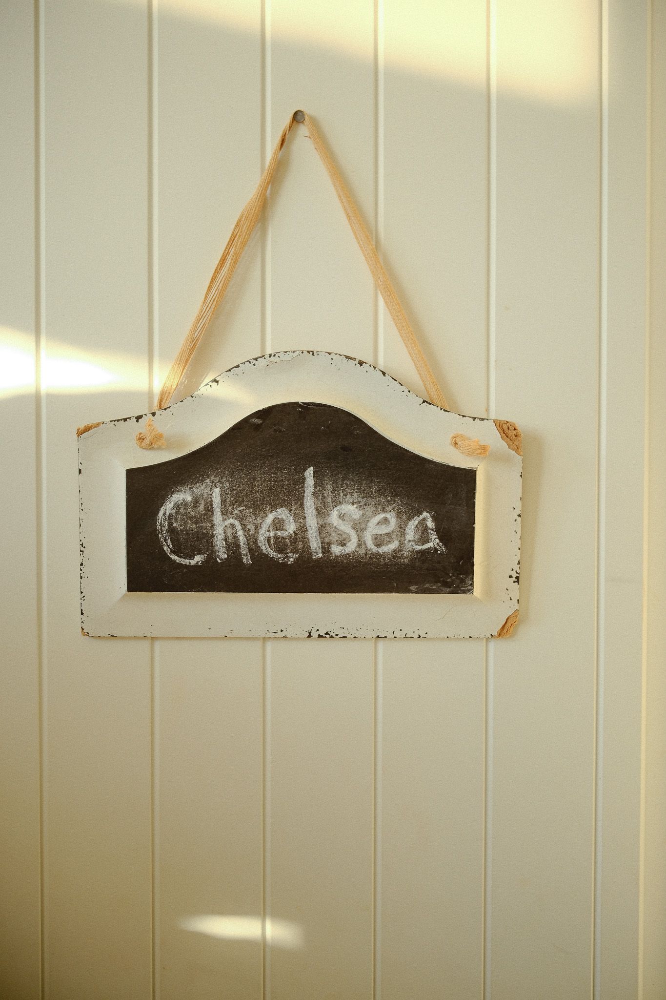 Rustic chalkboard sign with Chelsea written in white chalk hanging on white wood paneled wall.