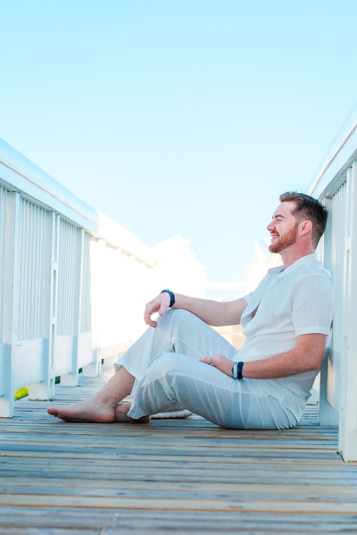 A smiling man in white linen outfit sits barefoot on a wooden boardwalk deck, relaxing against a white railing.