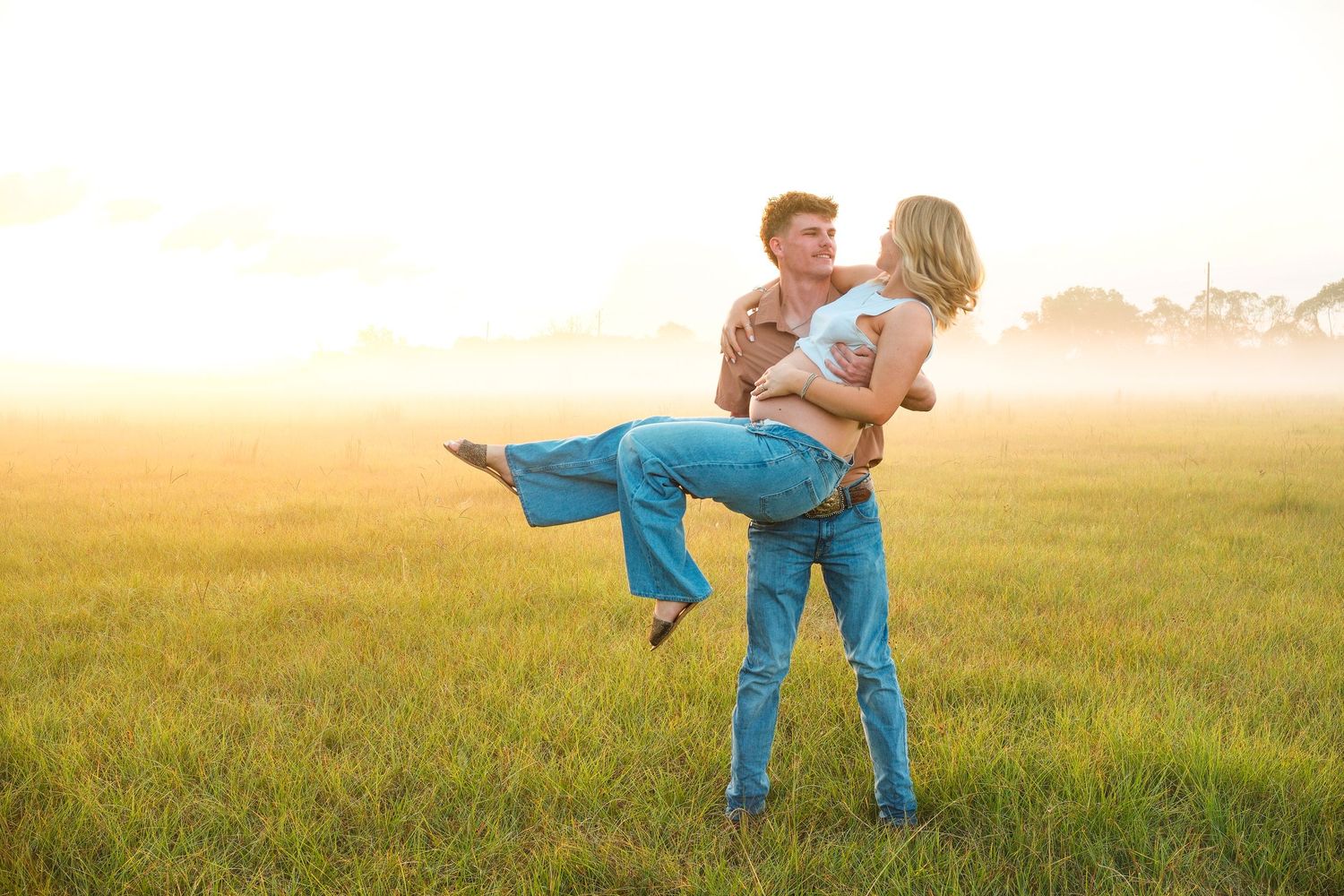 Couple dancing joyfully in a misty golden field at sunset, man lifting woman in jeans and boots.