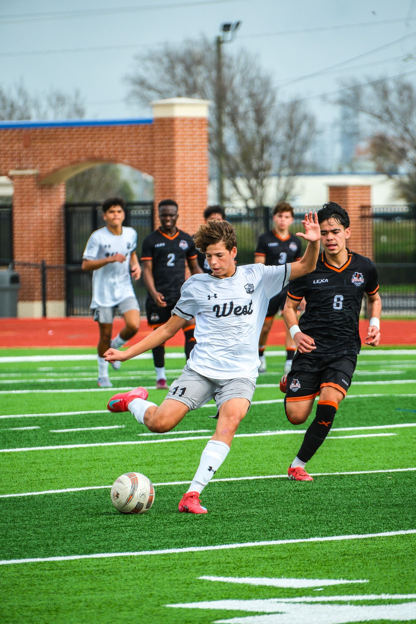 West high school soccer player in white uniform kicking the ball during a competitive match on turf.