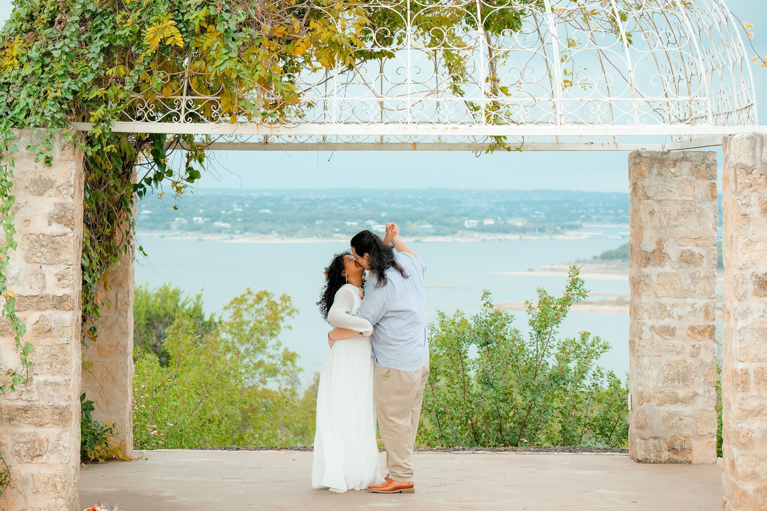 A couple shares a romantic kiss under a stone archway with a scenic lake view in the background.