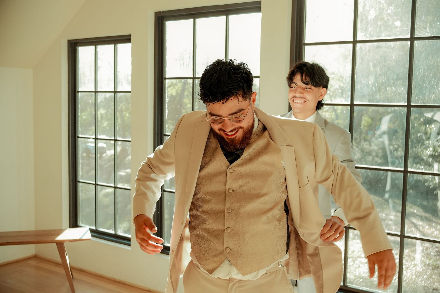 Two men in beige suits laugh together in a bright room with large windows during a wedding celebration.