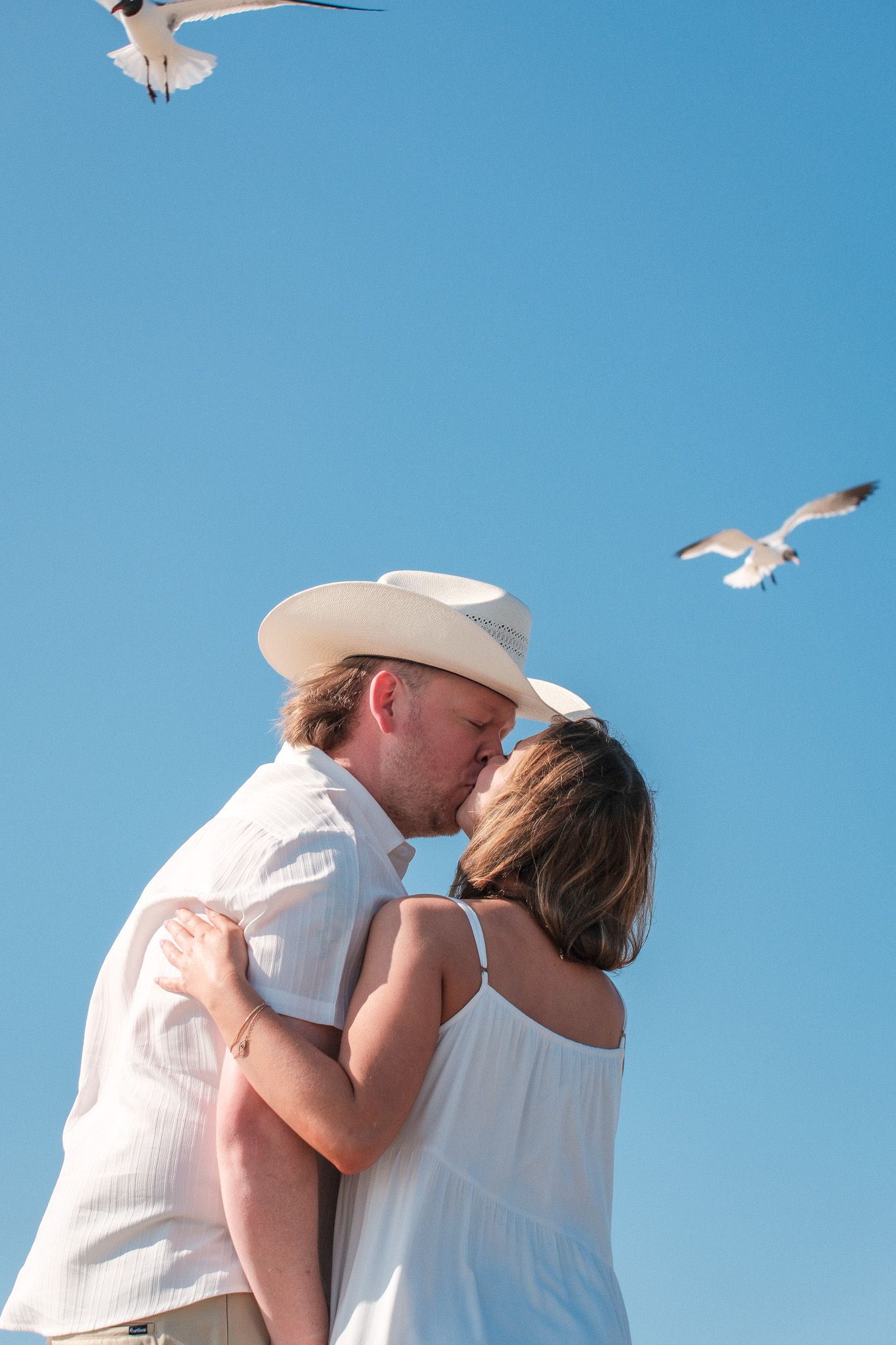 Couple sharing romantic beach kiss under blue sky with seagulls flying overhead.