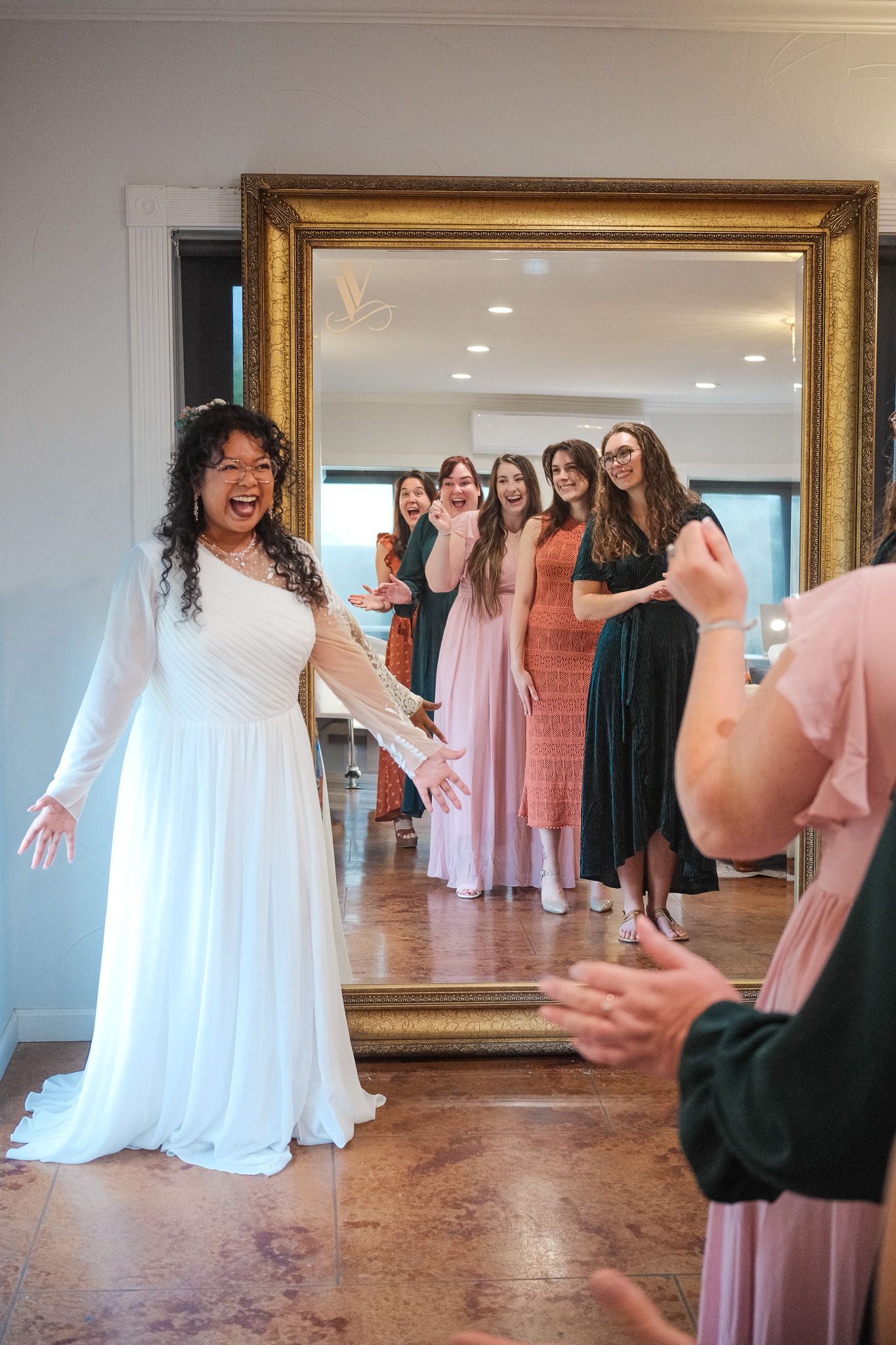 Bride in white gown laughs with bridesmaids reflected in ornate gold mirror during wedding preparations.