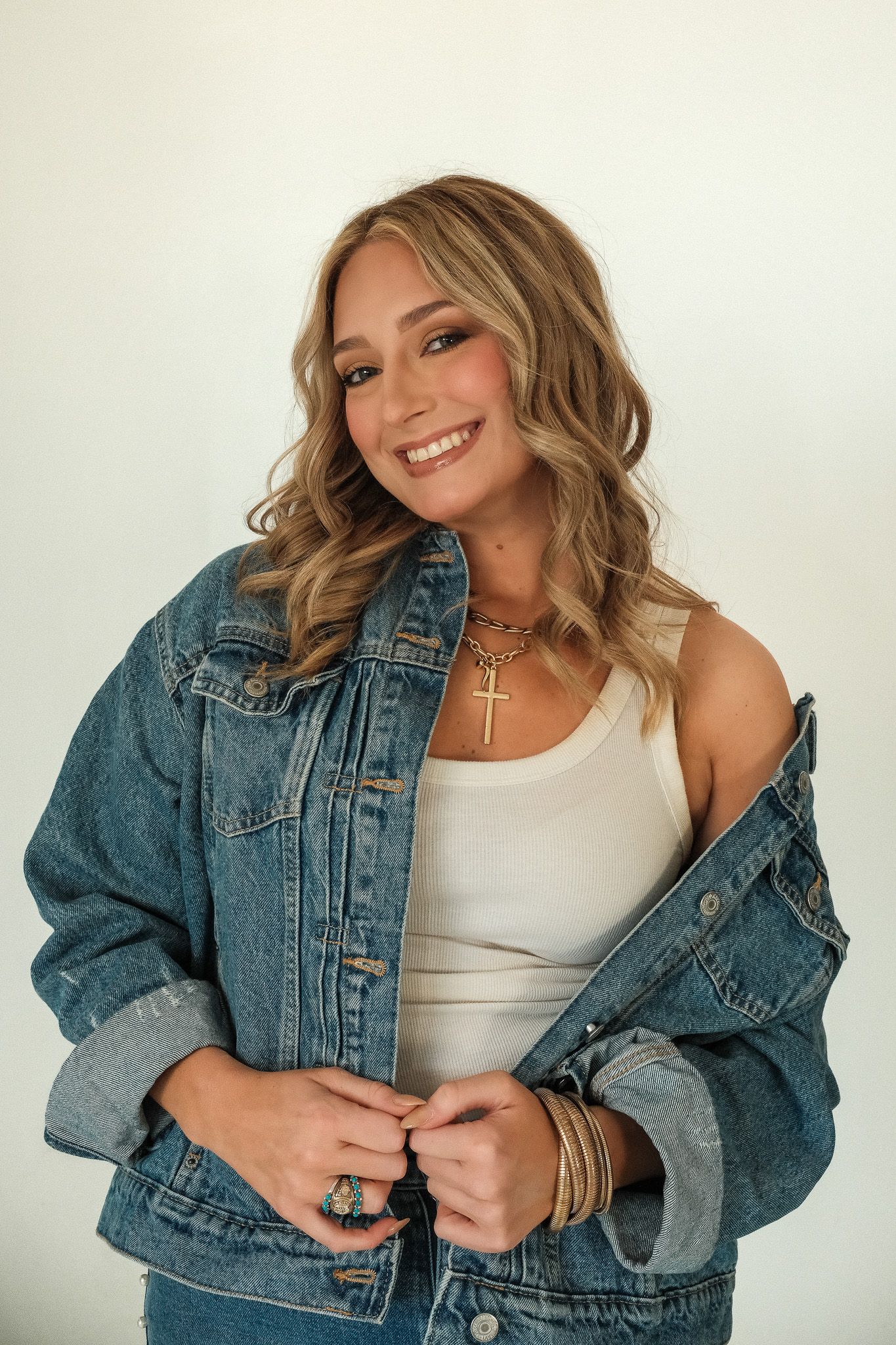 Smiling woman in a denim jacket over a white tank top with gold necklaces and bracelets.