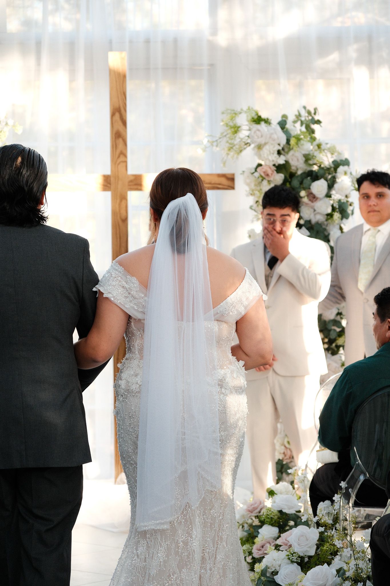 Emotional groom watches bride walk down aisle toward floral arch and wooden cross at wedding ceremony.